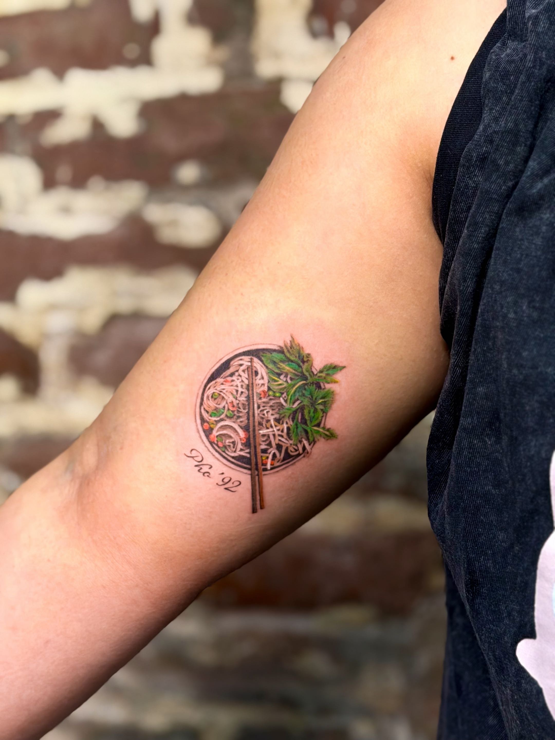 A person's upper arm with a colorful tattoo of a tree, circle, and text, with part of their black shirt visible and a blurred background of a brick wall.