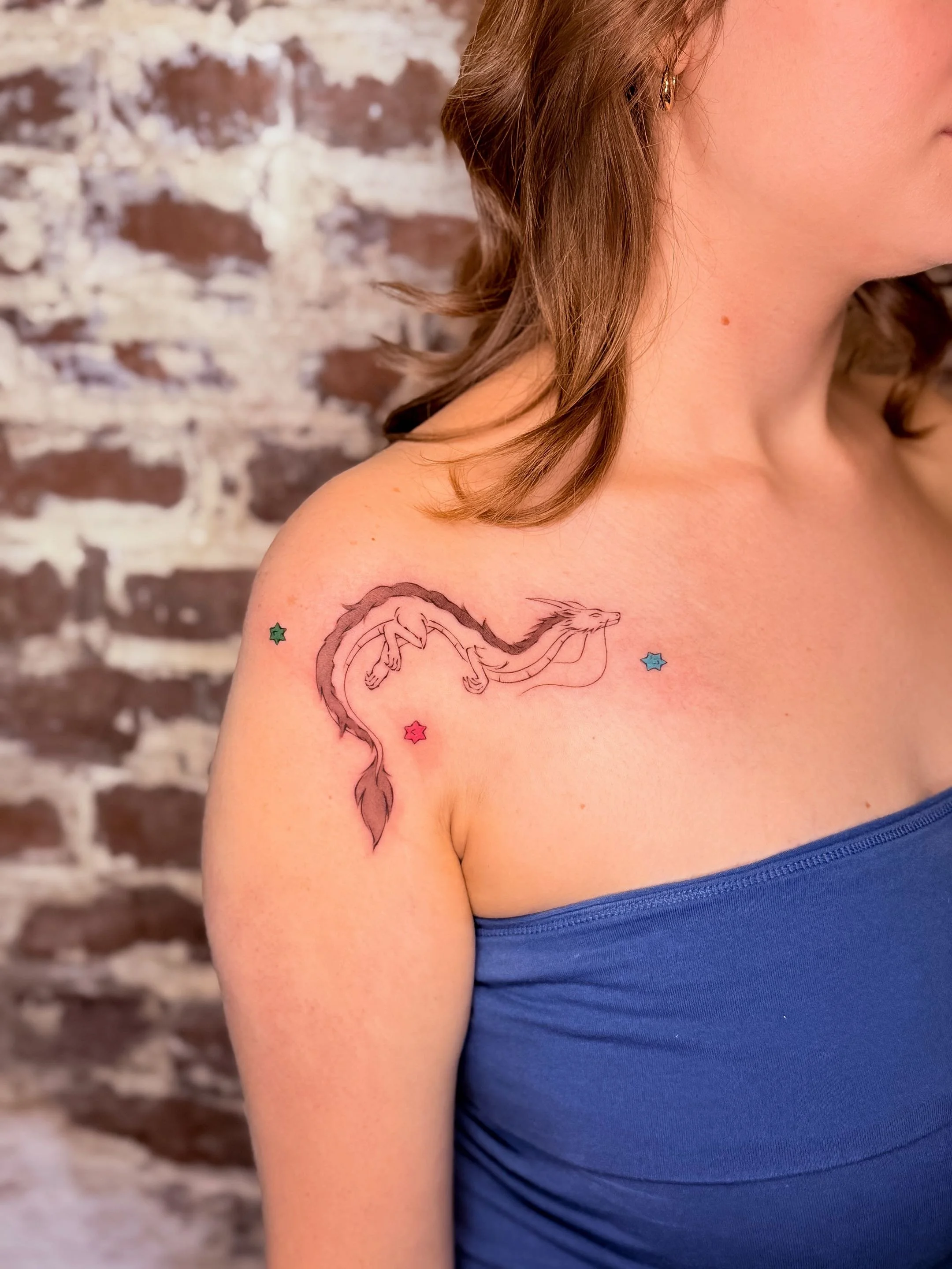Close-up of a woman's shoulder with a colorful dragon tattoo, wearing a blue top, with a brick wall in the background.