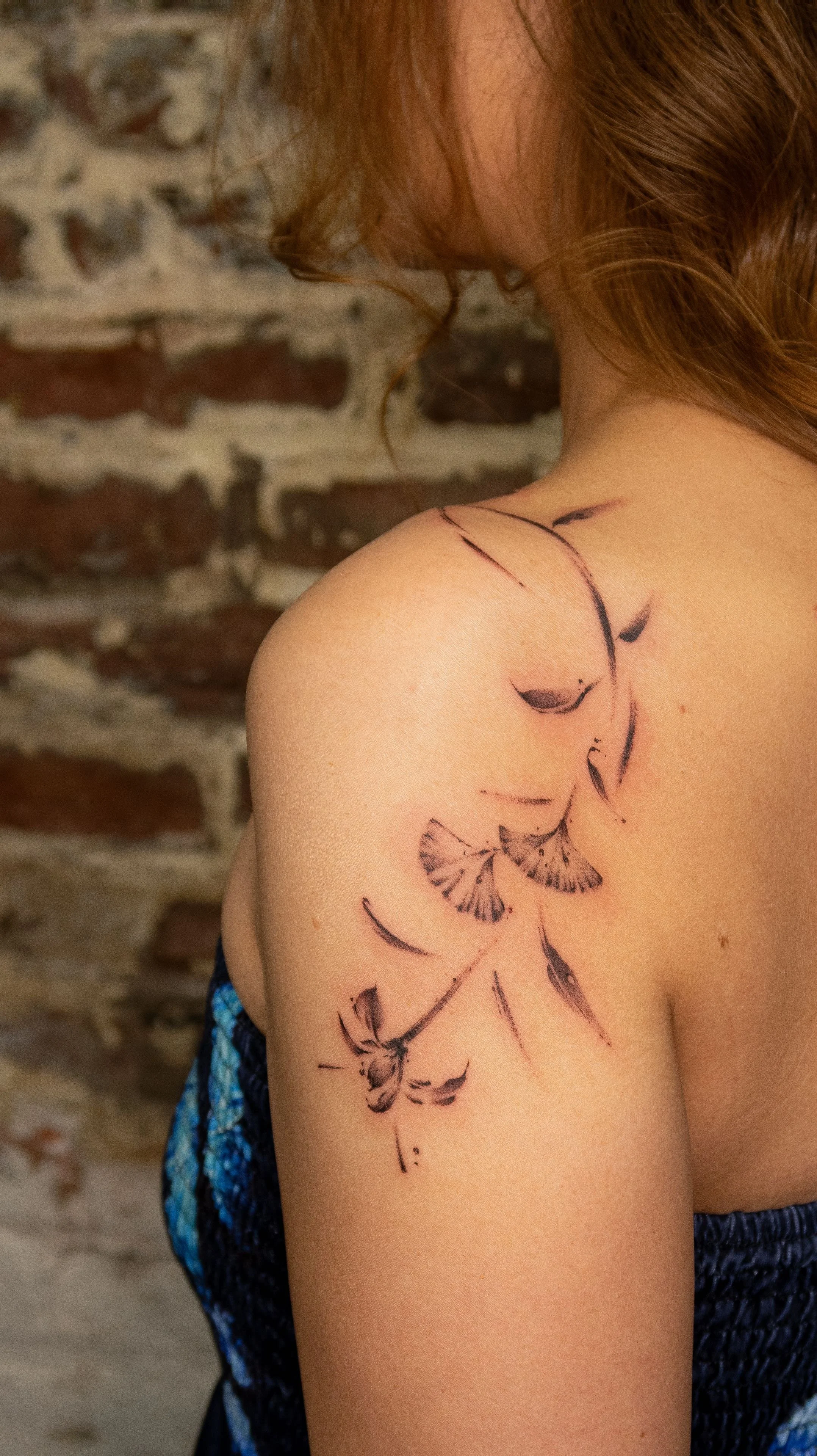 A woman with red hair showing her shoulder and upper arm, which has a black ink tattoo of a floral design and abstract pattern, with a brick wall background.
