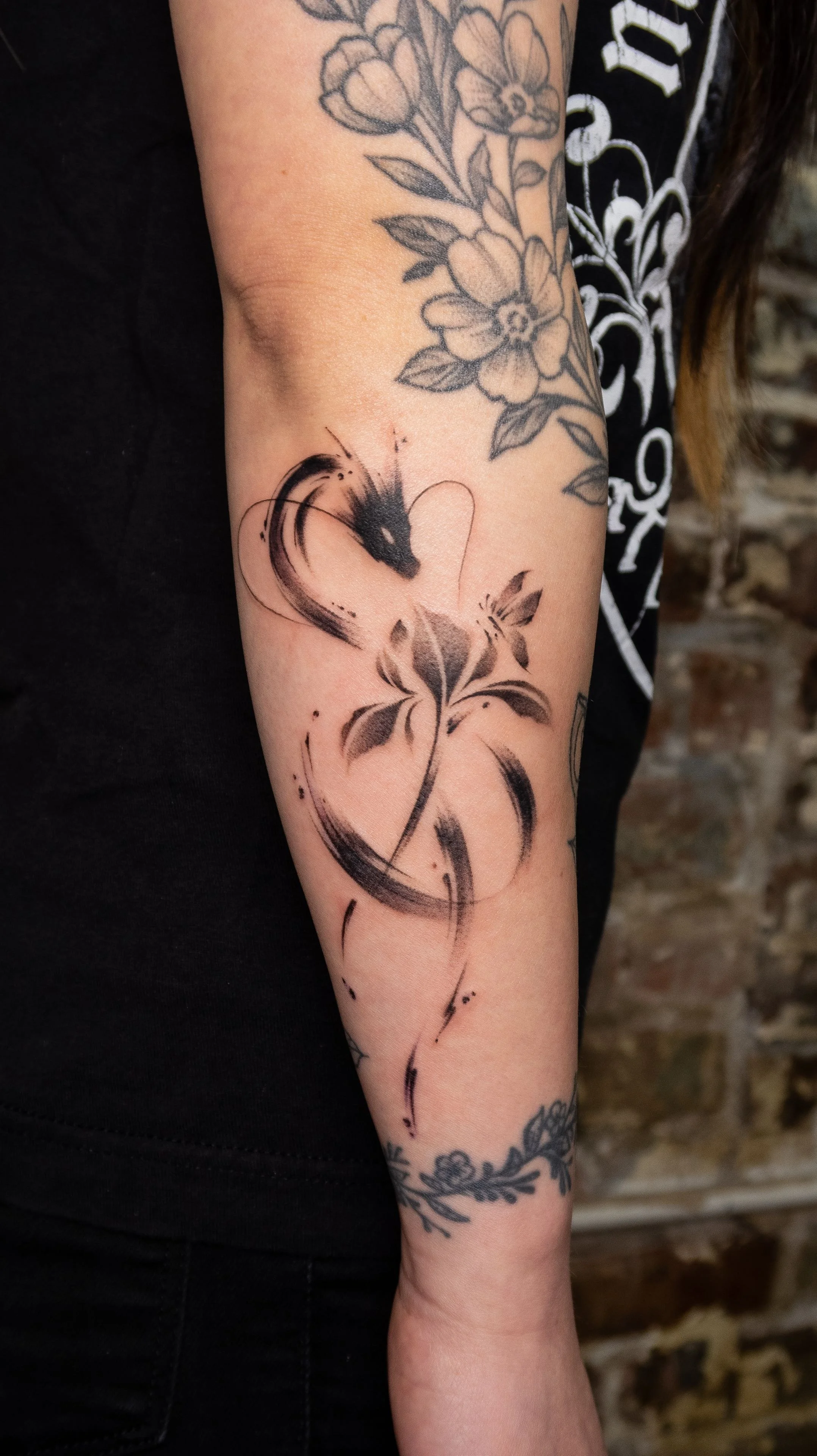 Tattoo of a black ink abstract bird with a long curved tail on the person's forearm, surrounded by floral tattoos on the upper arm.