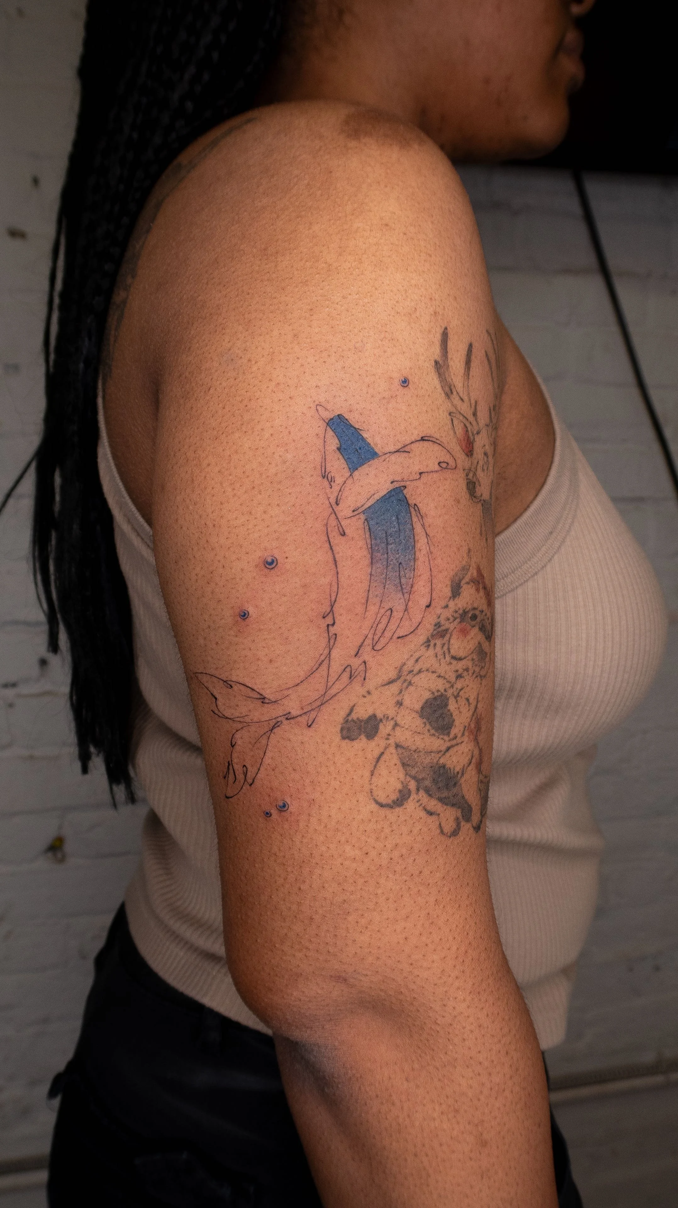 A woman with dreadlocks and freckles wearing a beige tank top shows her shoulder and upper arm with tattoos of a blue butterfly, a lion, and small blue dots.