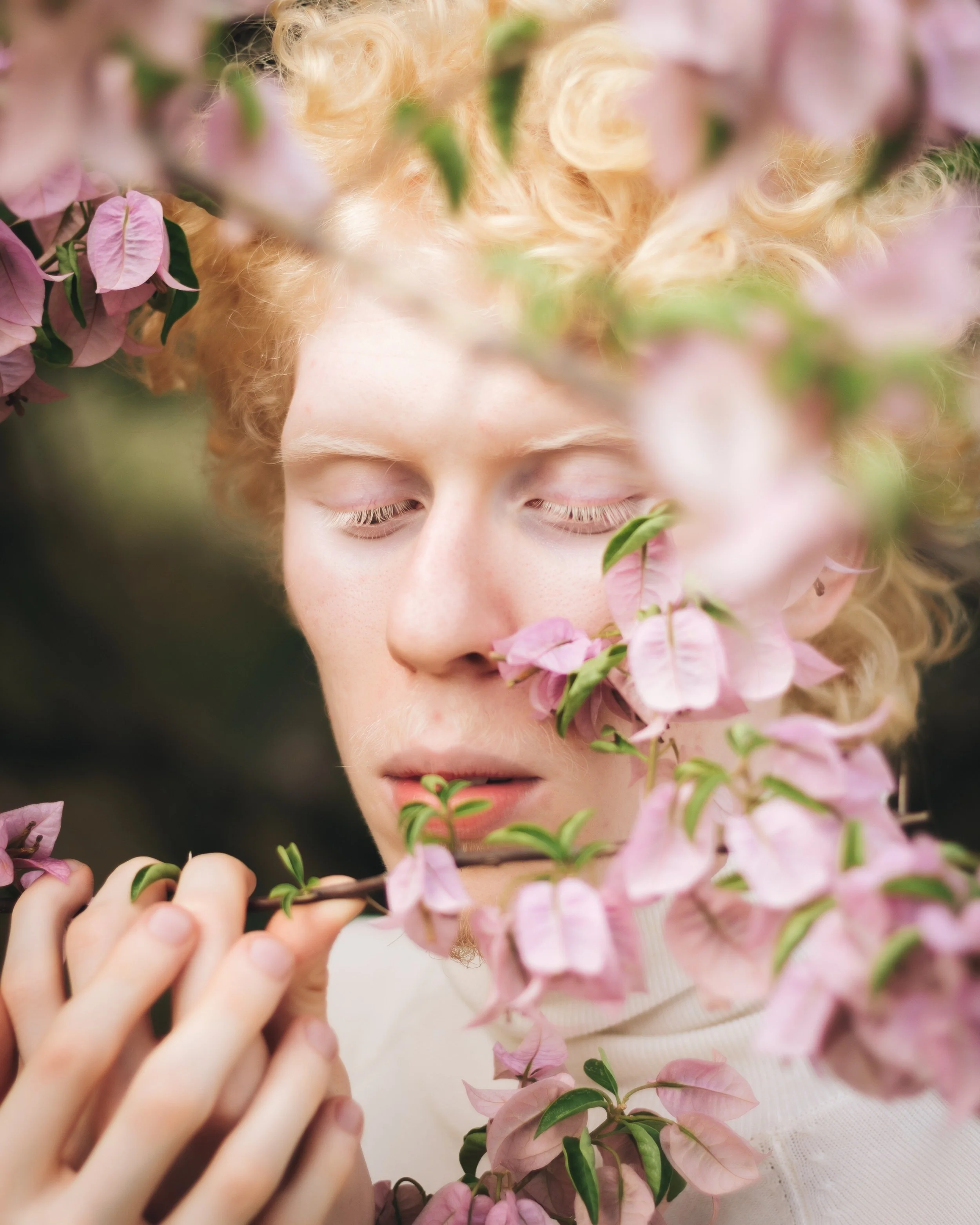 Close-up of a person with light skin and curly blonde hair amidst pink and green foliage, eyes closed, gently holding a branch of pink flowers close to their face.