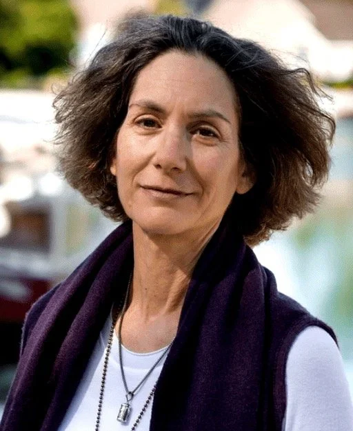 A woman with short, curly brown hair wearing a dark scarf and a white top, outdoors with blurred boats in the background.
