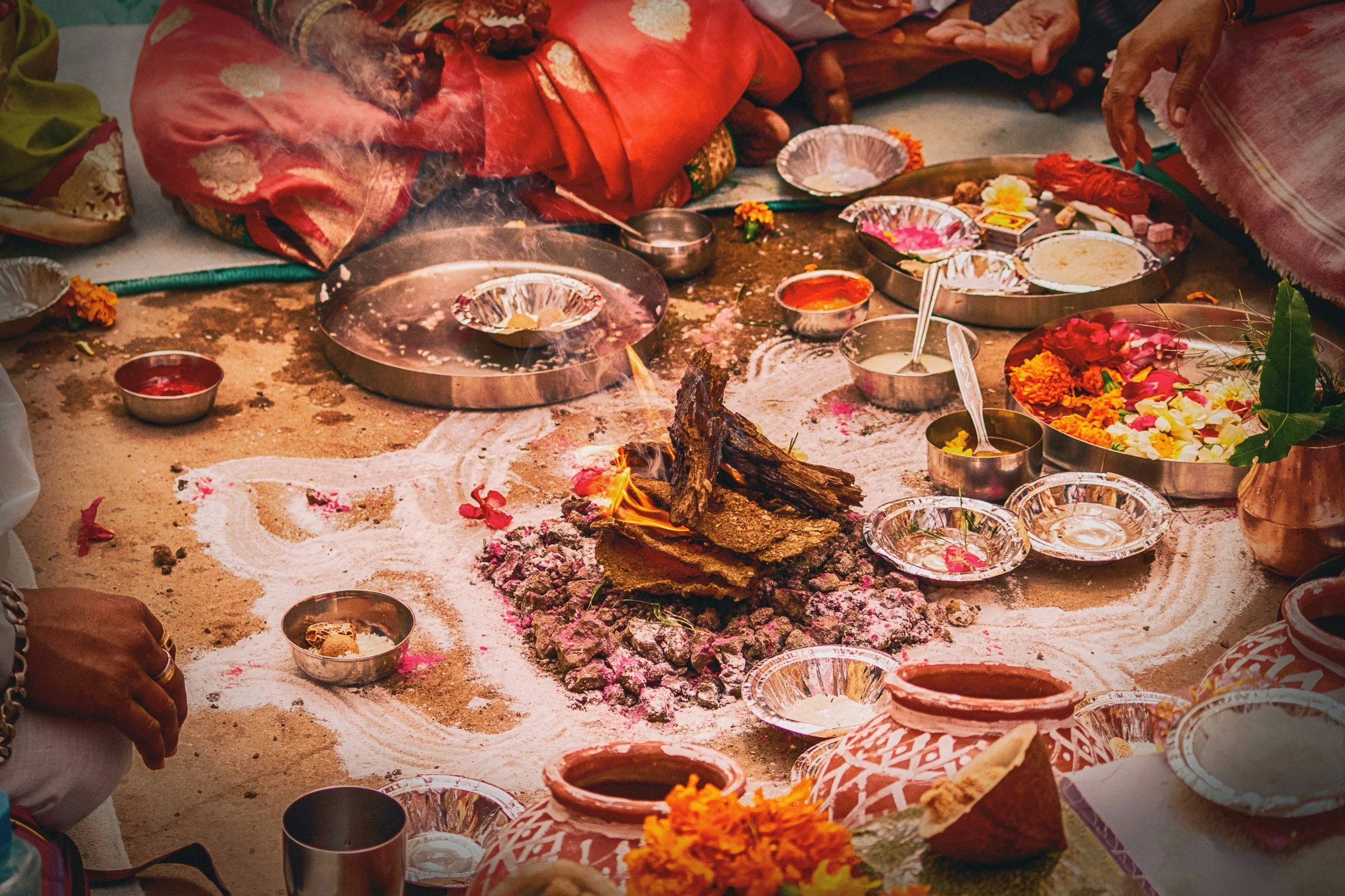 A traditional Hindu or Indian religious ceremony with an altar on the ground. The setup includes burning logs, offerings, flowers, and various ceremonial items arranged around a fire.