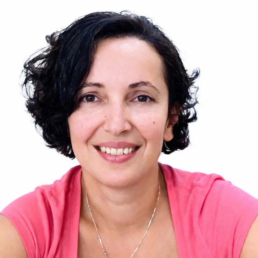 Close-up of a woman with short black curly hair smiling, wearing a pink top and a silver necklace, against a white background.