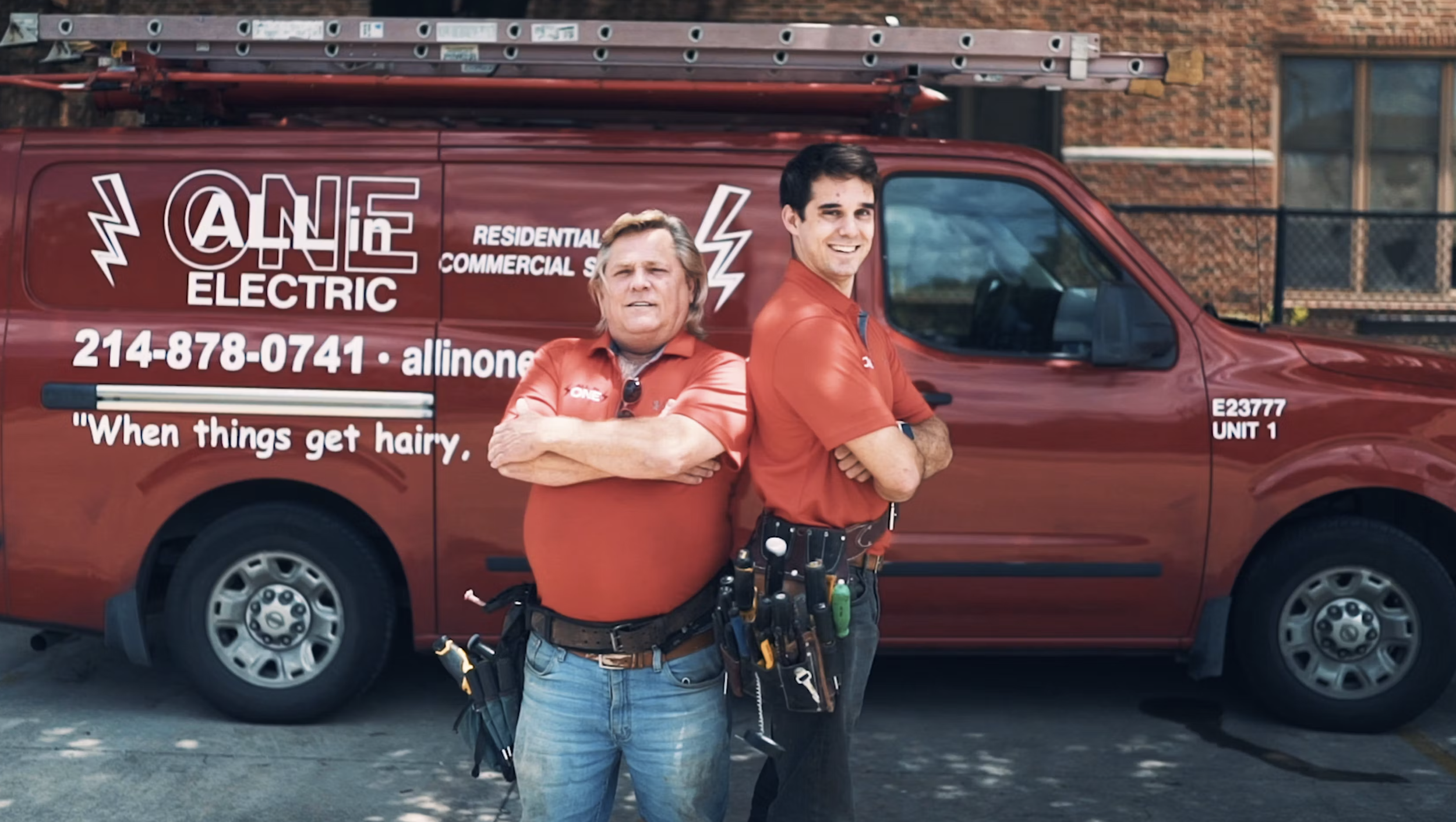 Dual Master Electrician father-son team standing in front of a red service van with company branding, both with crossed arms, wearing red shirts and tool belts.