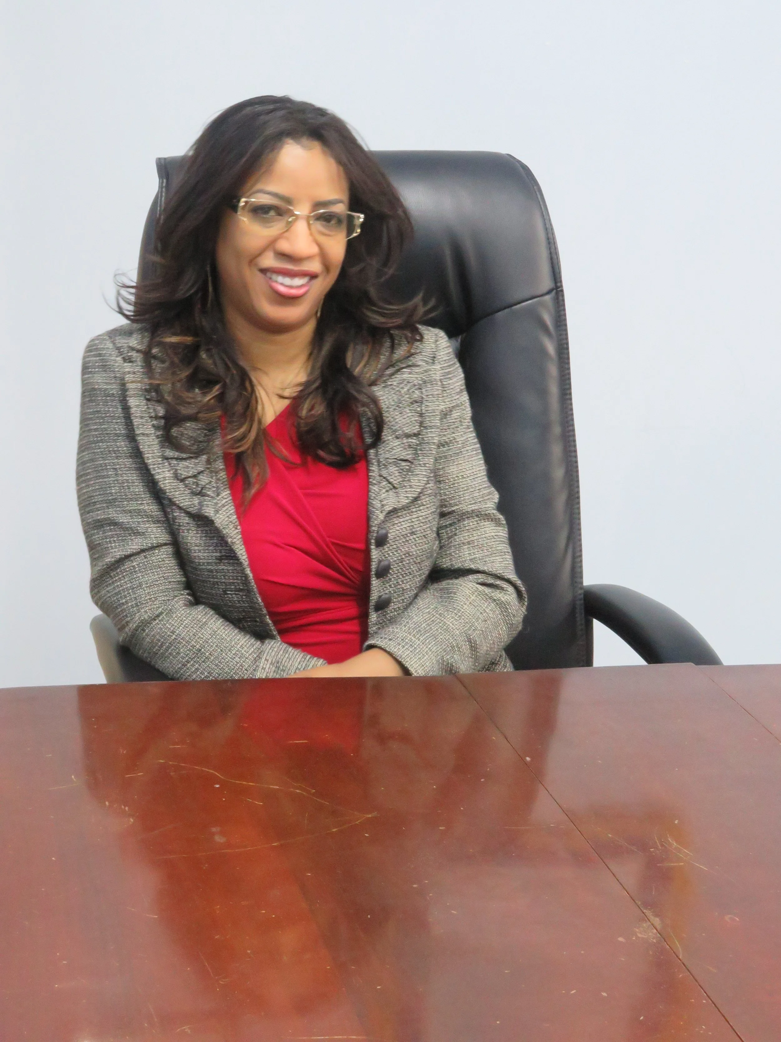 A professional woman with long dark hair, glasses, and a blazer, sitting on a black office chair at a wooden conference table.