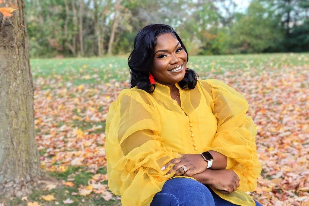 A woman in a yellow blouse sitting outdoors in a park during autumn, surrounded by fallen leaves and trees.