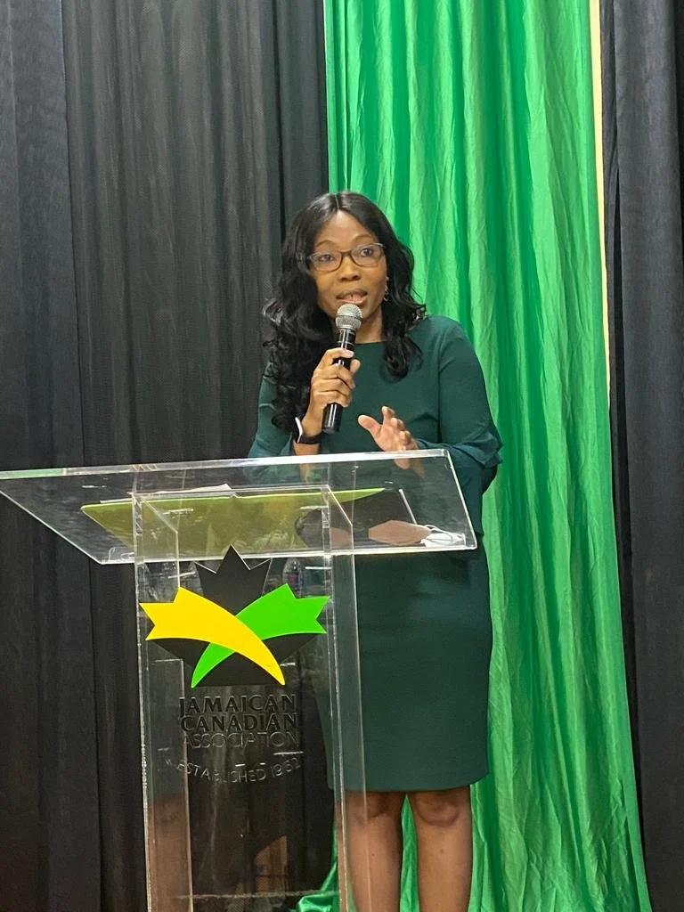 A woman in a green dress speaking into a microphone at a podium with the Jamaica Canadian Association logo, in front of black and green curtains.