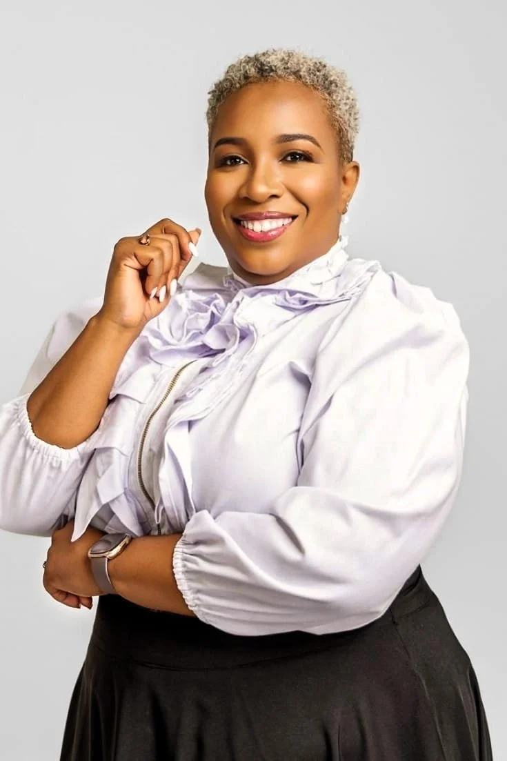 A smiling woman with short, curly, blonde hair wearing a white blouse with ruffled details and a black skirt, standing against a plain background.