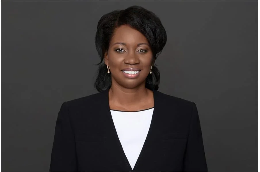 Portrait of a smiling Black woman with short black hair, wearing a black blazer over a white top, against a gray background.