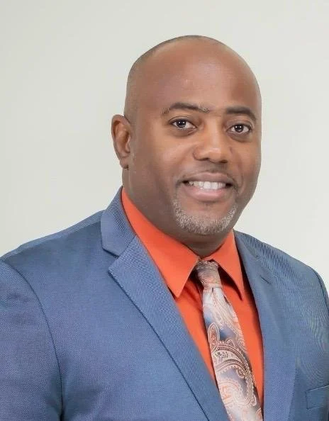 A man in a blue suit, orange shirt, and paisley tie smiling at the camera against a plain white background.
