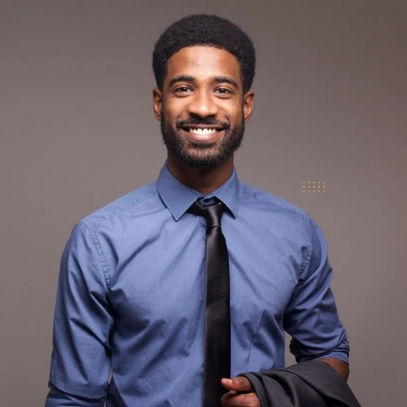 A smiling man with a beard and short hair wearing a blue dress shirt, black tie, and holding a black jacket, standing against a plain gray background.