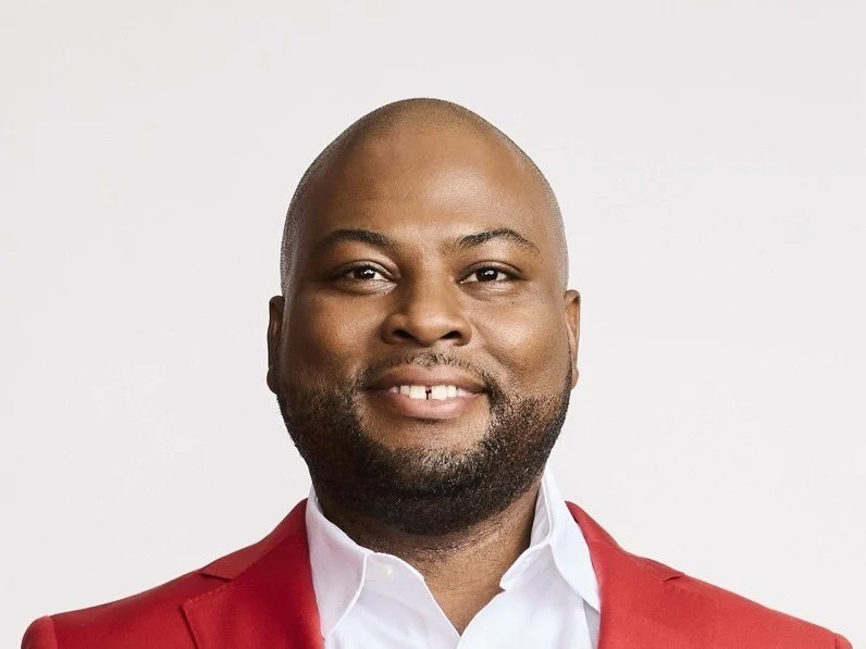 A man with a shaved head and beard wearing a red blazer and white shirt smiling against a light background.