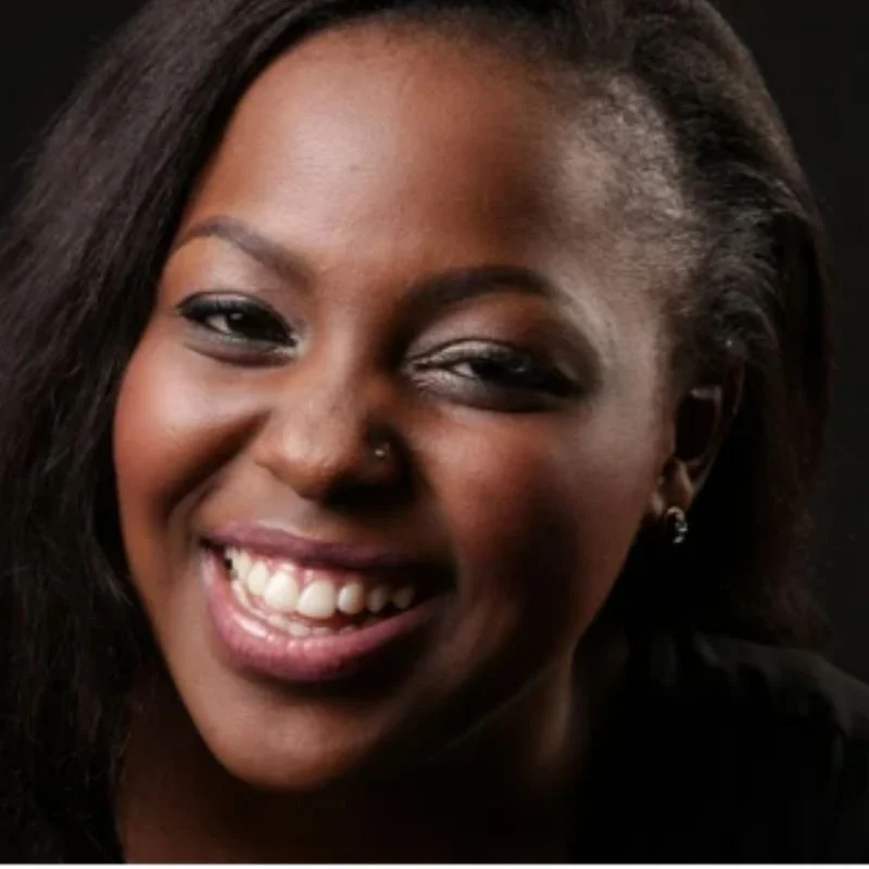 A close-up portrait of a smiling Black woman with dark hair, wearing a nose stud and earrings, against a dark background.