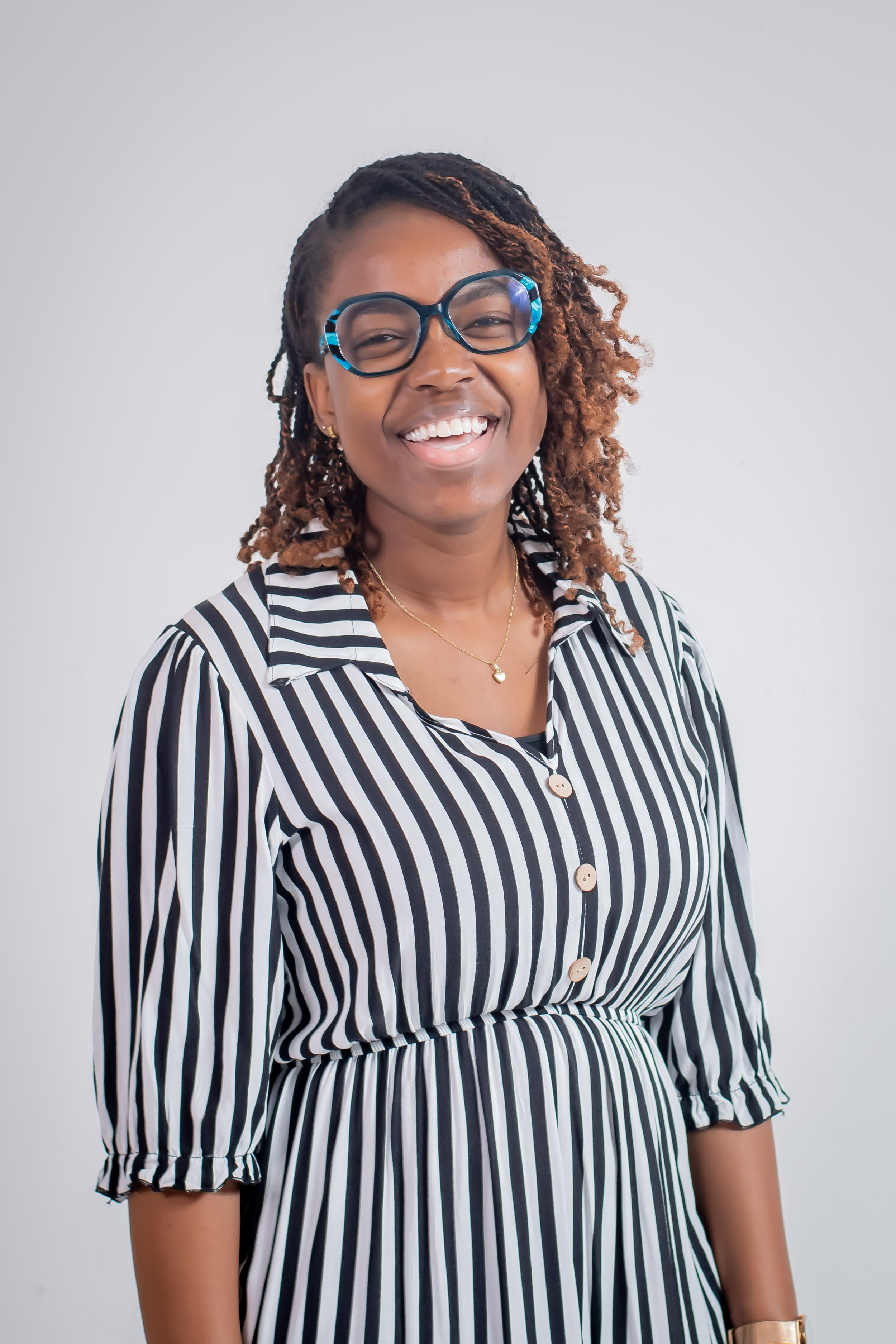 A smiling woman with glasses and curly hair, wearing a black and white striped dress with button details, standing against a plain white background.