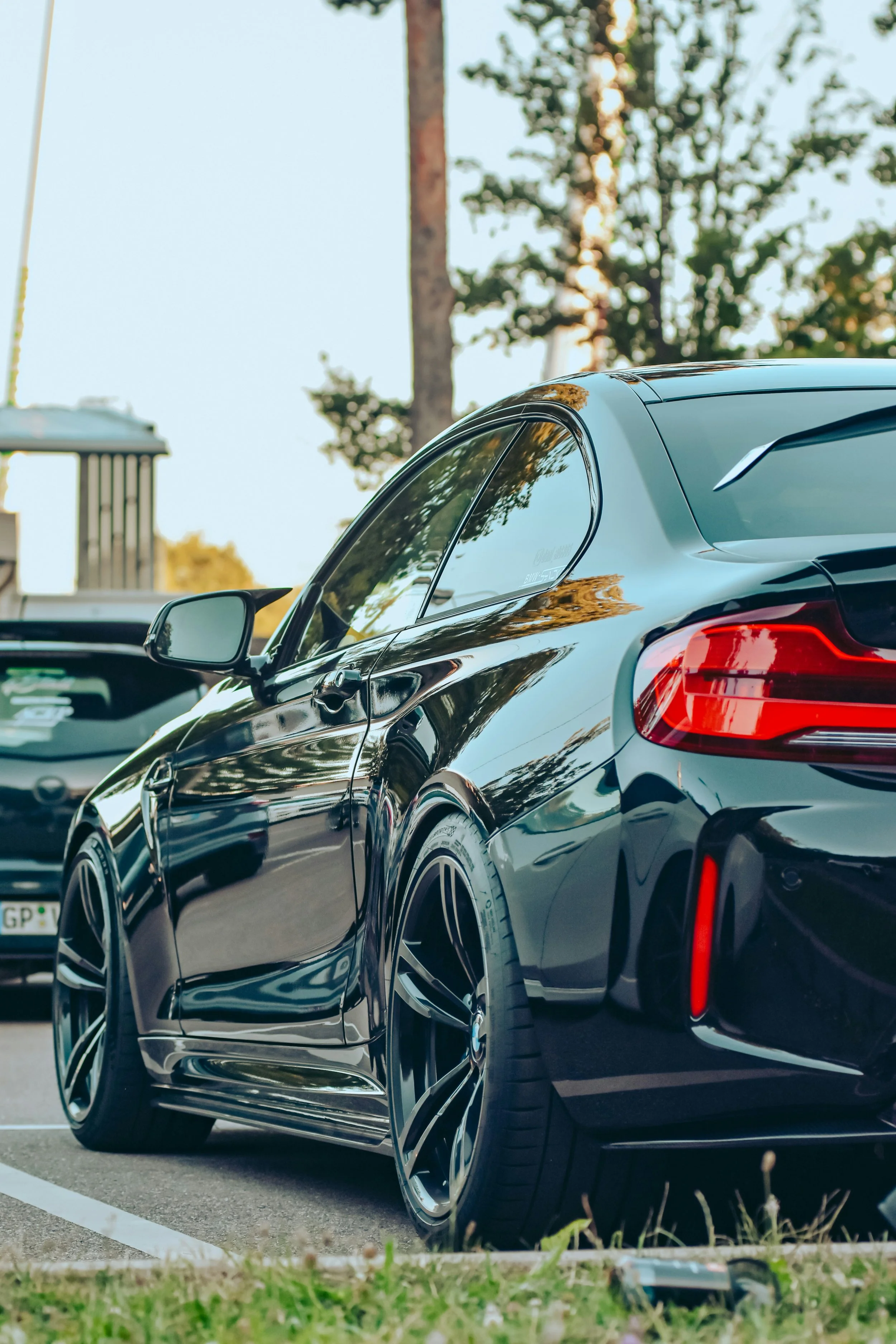 A black sports car parked in a parking lot with trees and another vehicle in the background.