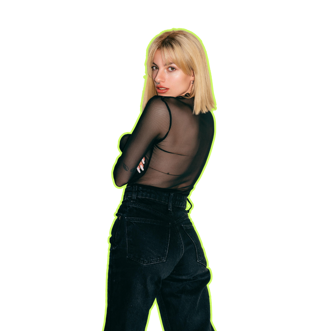 A young woman with blonde hair and a septum piercing wearing a black sheer long-sleeve top and high-waisted black jeans, posing with her back slightly turned and looking over her shoulder.