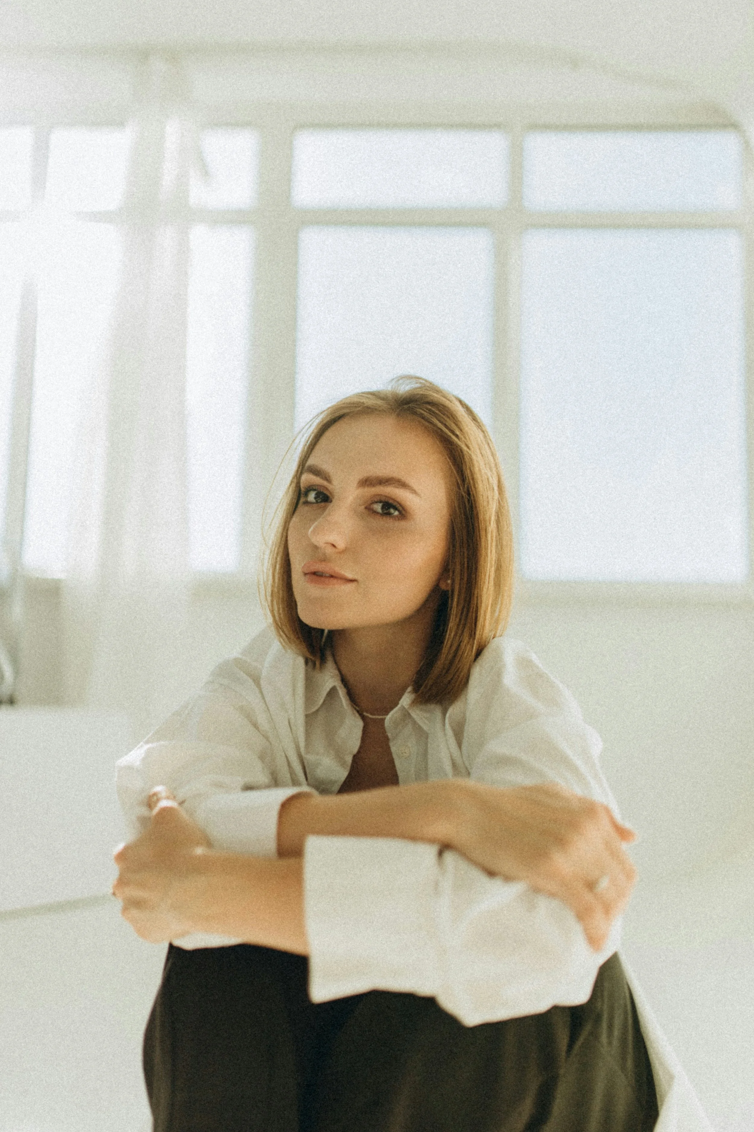 A woman with shoulder-length red hair and a neutral expression sitting in front of a large window with white curtains, wearing a white shirt and dark pants.