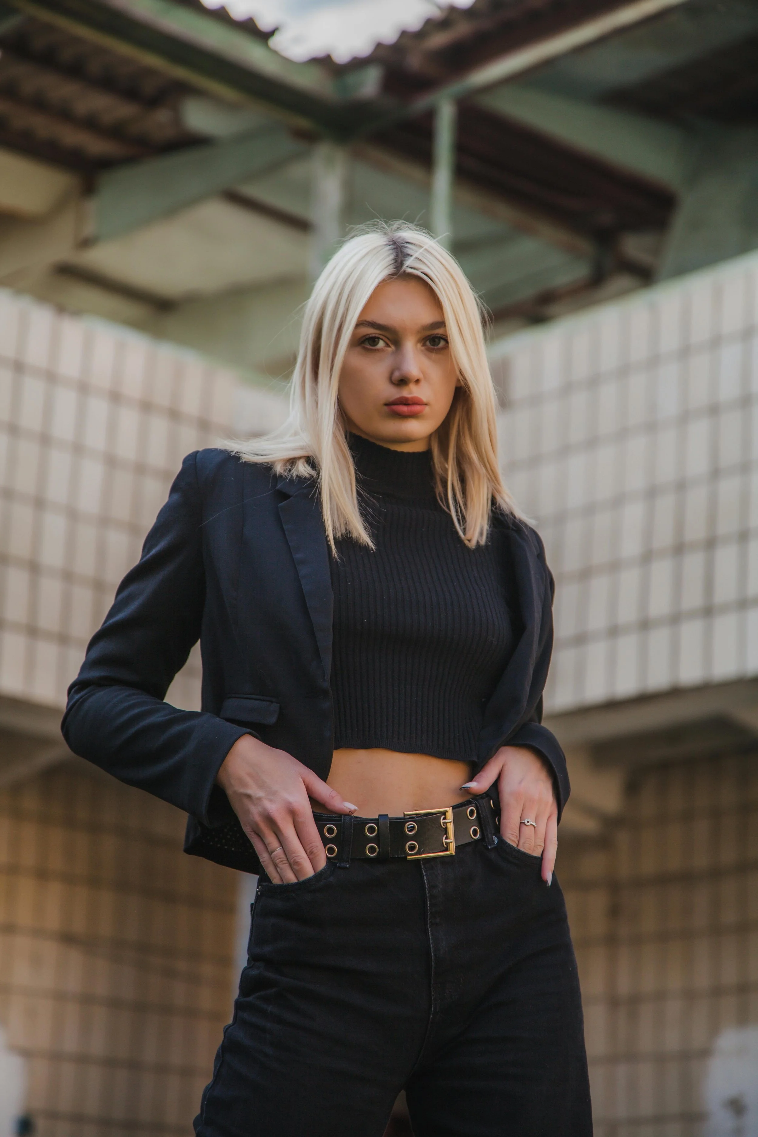A woman with blonde hair wearing a black turtleneck, black blazer, and high-waisted black jeans, posing confidently outdoors in front of a beige brick wall and a partially covered structure.