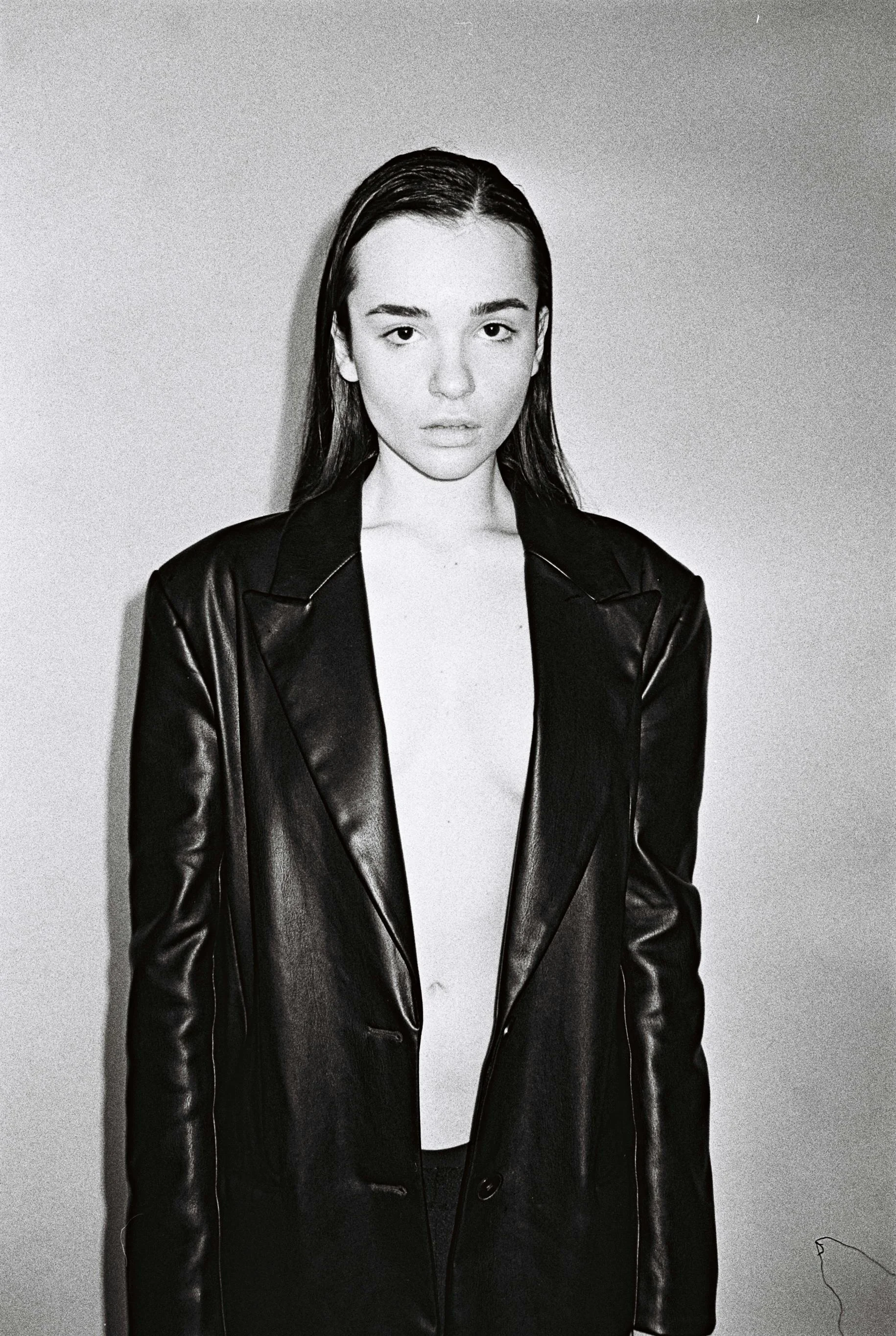 Black and white photo of a young woman with wet hair wearing a long leather coat, standing against a plain background.