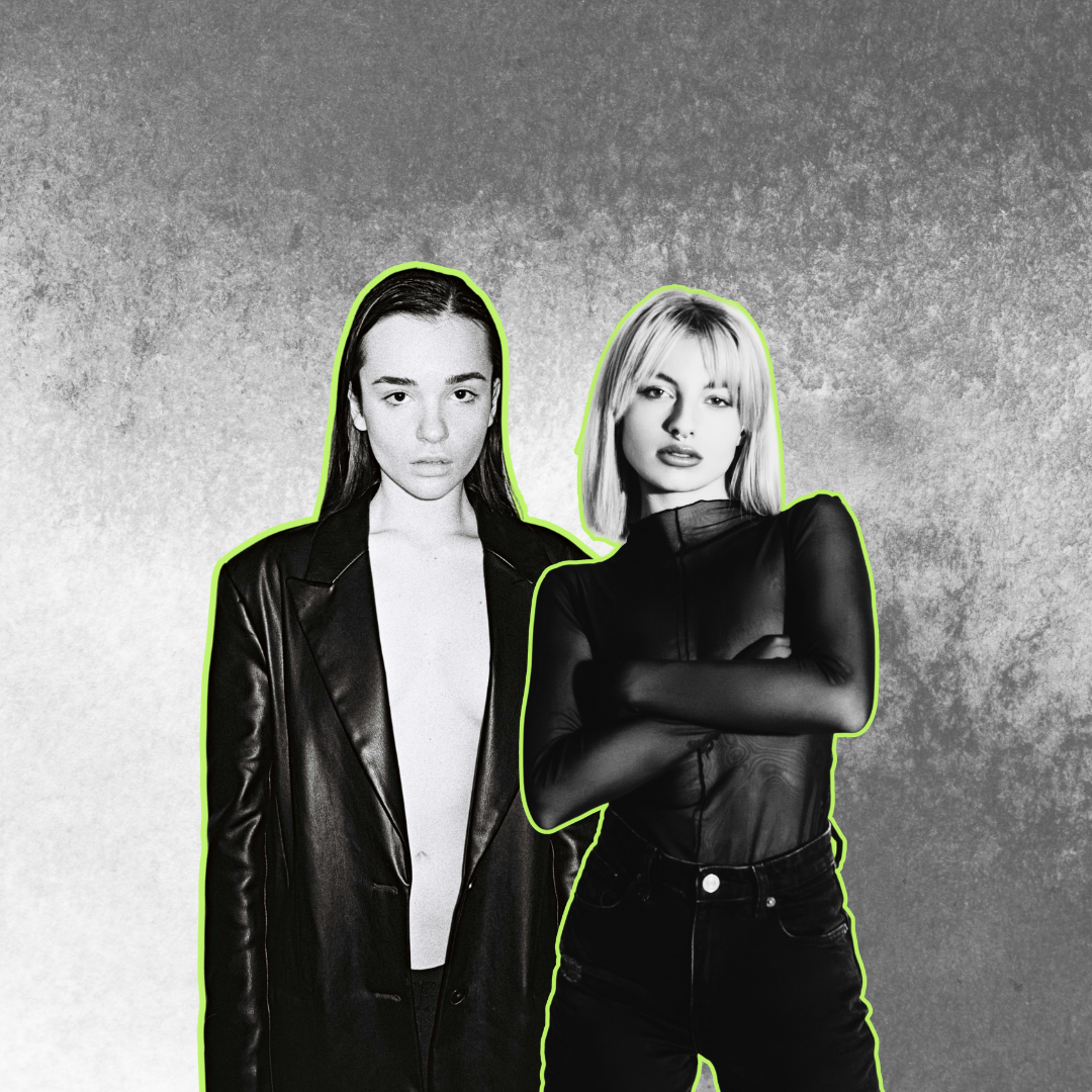 Black and white photo of two women standing side by side against a textured background, with neon green outlines around their figures. The woman on the left has long dark hair and is wearing an open leather jacket without a shirt underneath, while the woman on the right has short blond hair and is wearing a sheer long-sleeve top with crossed arms.