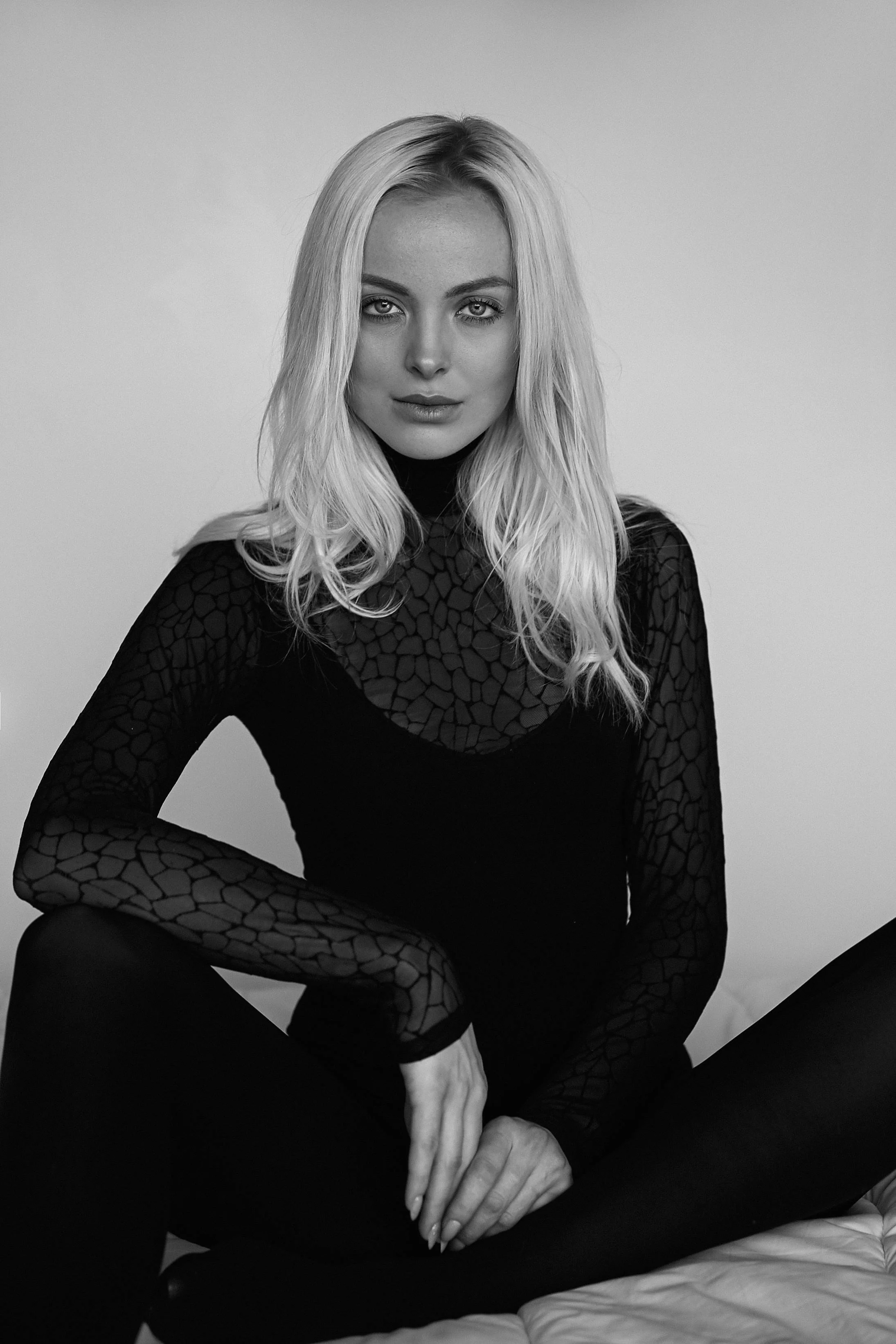 A black and white portrait of a woman with long blonde hair, seated on a bed, wearing a dark patterned top and black pants, looking directly at the camera.