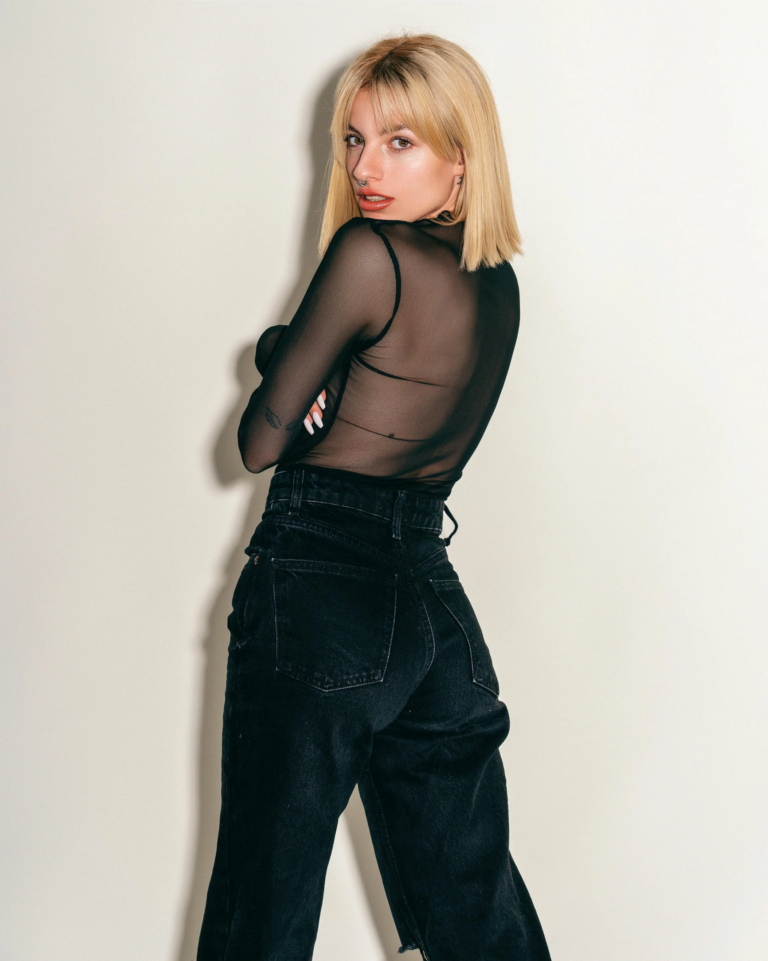 A young woman with blonde hair, wearing a black sheer top and high-waisted black jeans, standing against a plain white background.