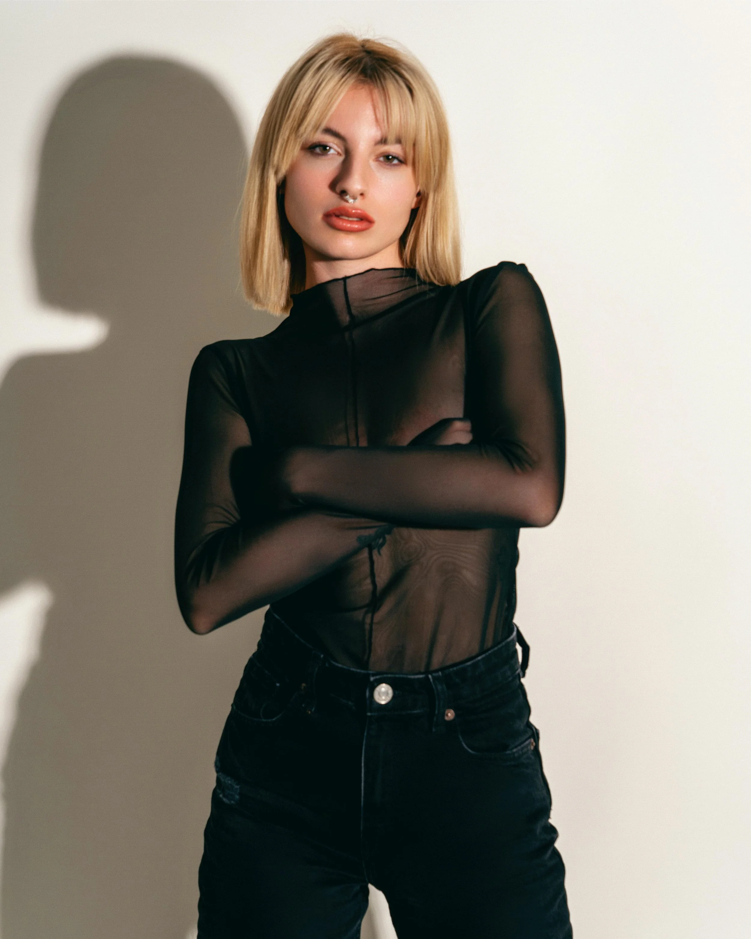 A woman with blonde hair wearing a sheer black long-sleeve top and black pants, standing against a white background with shadows.