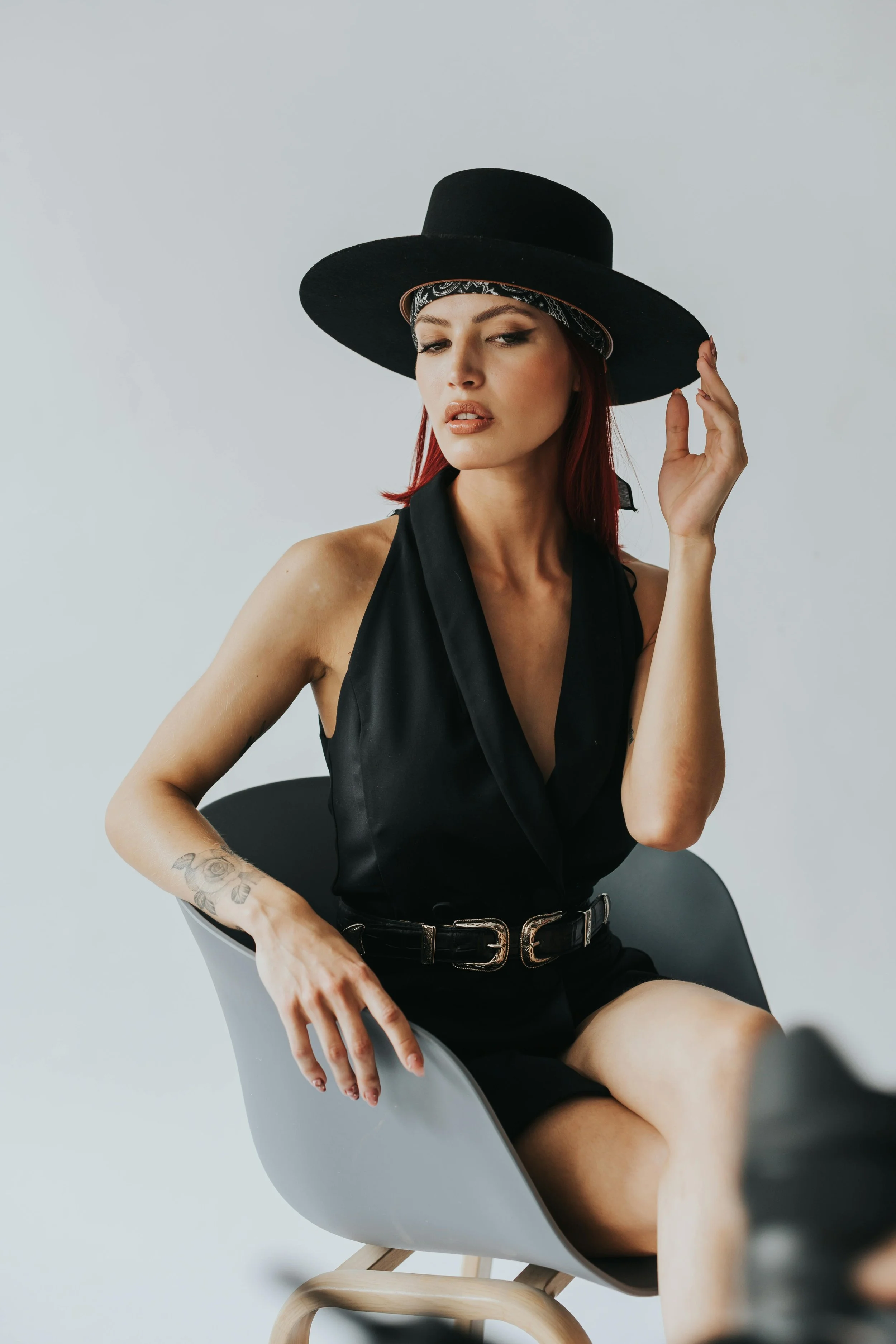 A woman with red hair and tattoos sits on a modern gray chair, wearing a black sleeveless top, a wide-brimmed black hat, and a patterned bandana.