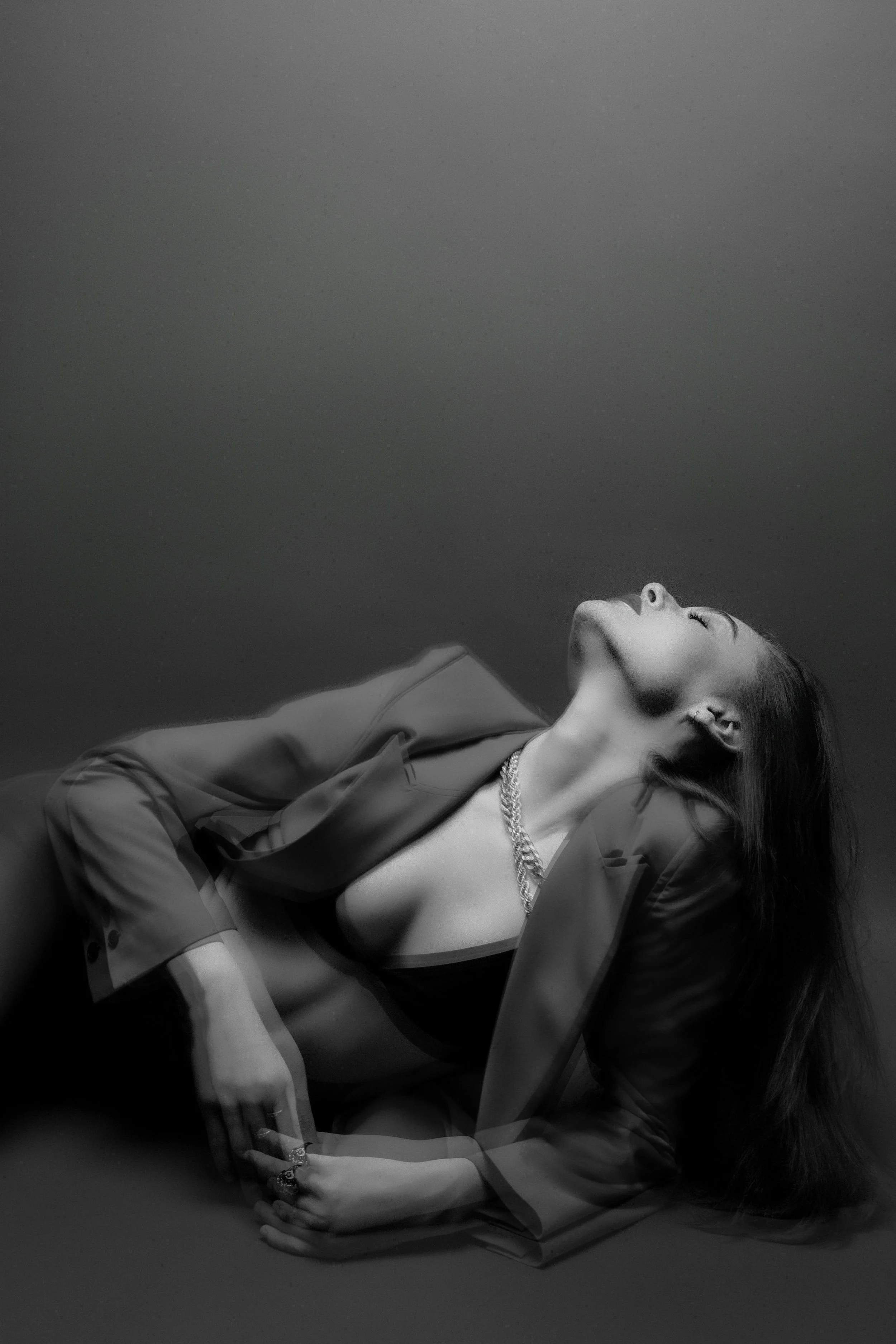 Black and white portrait of a woman lying on her side with her head tilted back, wearing a blazer and jewelry.