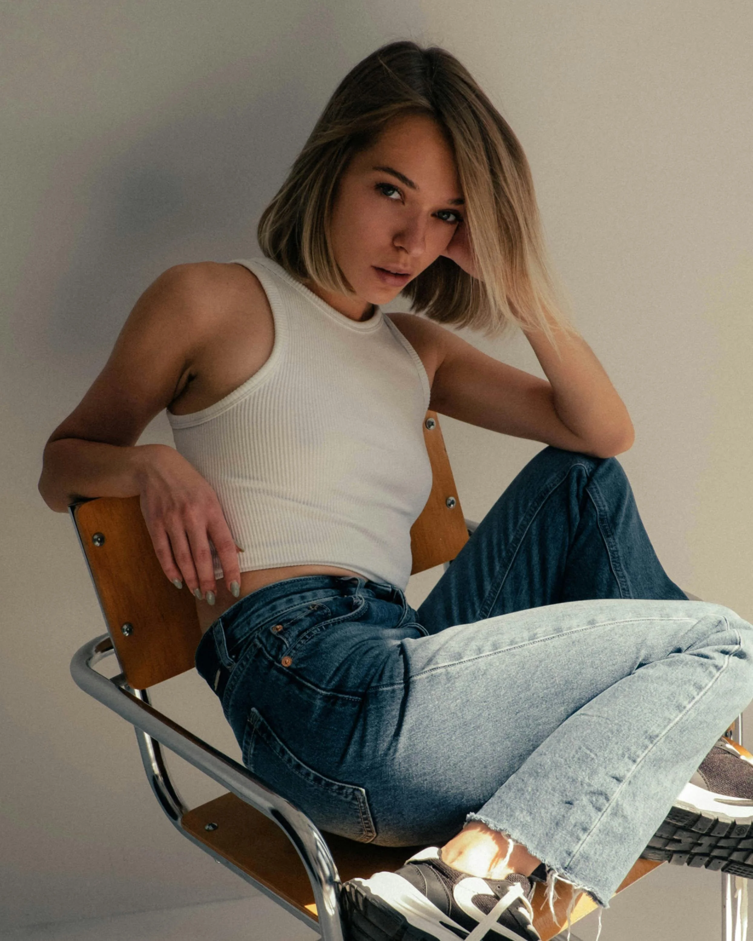 A young woman sitting on a wooden and metal chair, wearing a white sleeveless top, high-waisted jeans, and sneakers, with her head tilted and hand resting on her head.