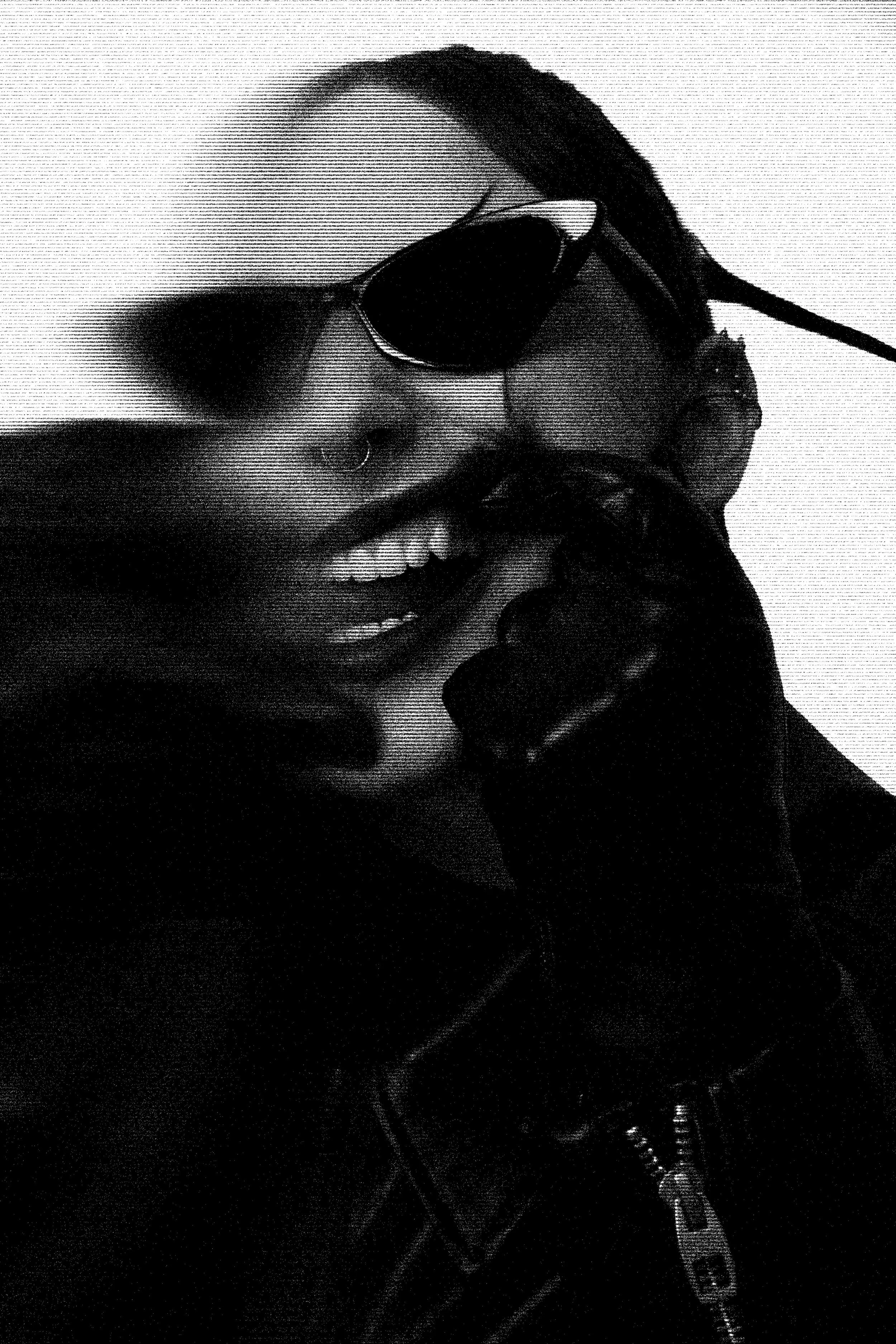 Black and white photo of a smiling woman wearing sunglasses, a nose ring, a leather jacket, and gloves, holding a telephone receiver to her ear.