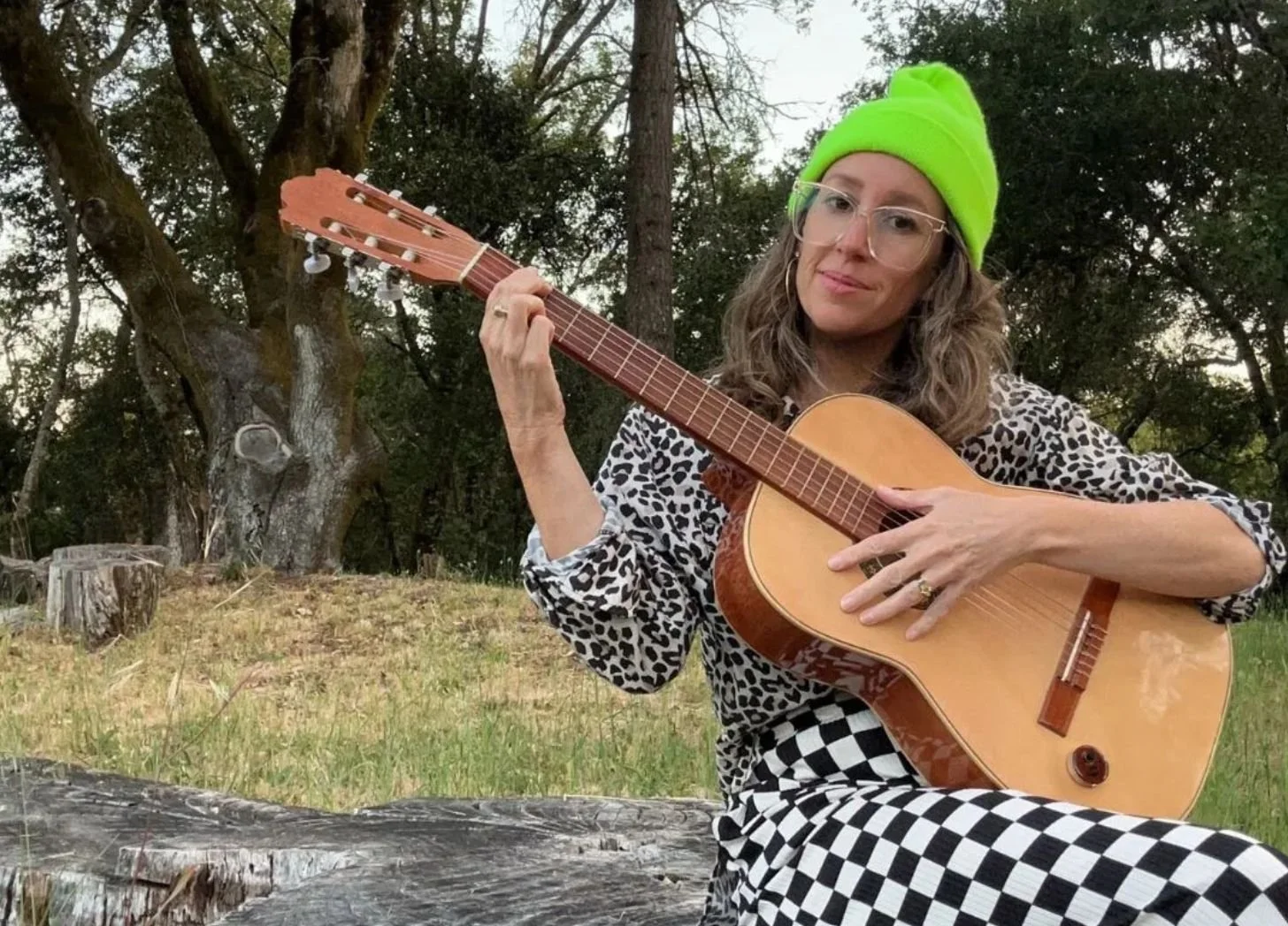 Little Door sitting outdoors playing an acoustic guitar, wearing a neon green beanie, glasses, a leopard print top, and checkered pants, with trees in the background.
