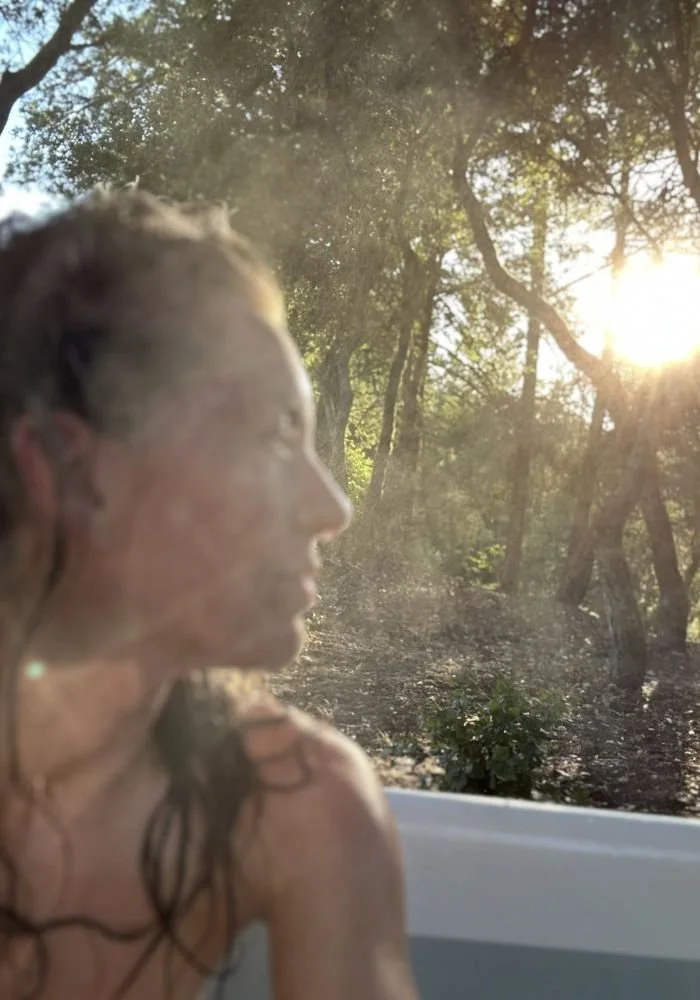 Little Door with wet hair is sitting in a bathtub outdoors with trees and sunlight in the background.