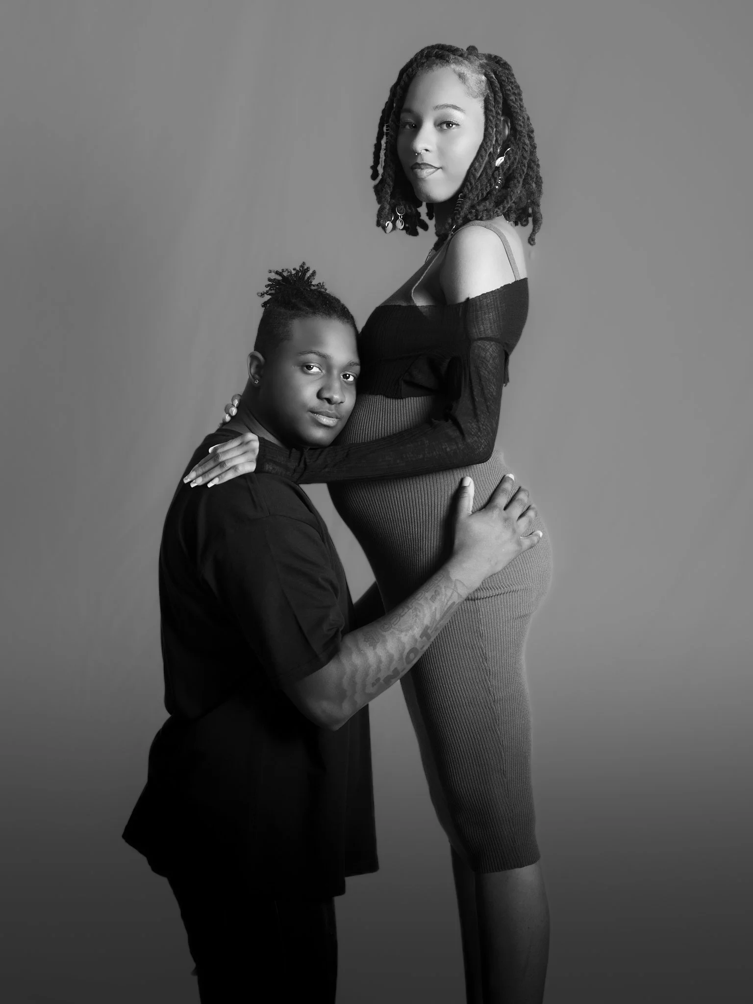 Black and white image of a couple, with the man kneeling and holding a pregnant woman's belly, both looking at the camera.