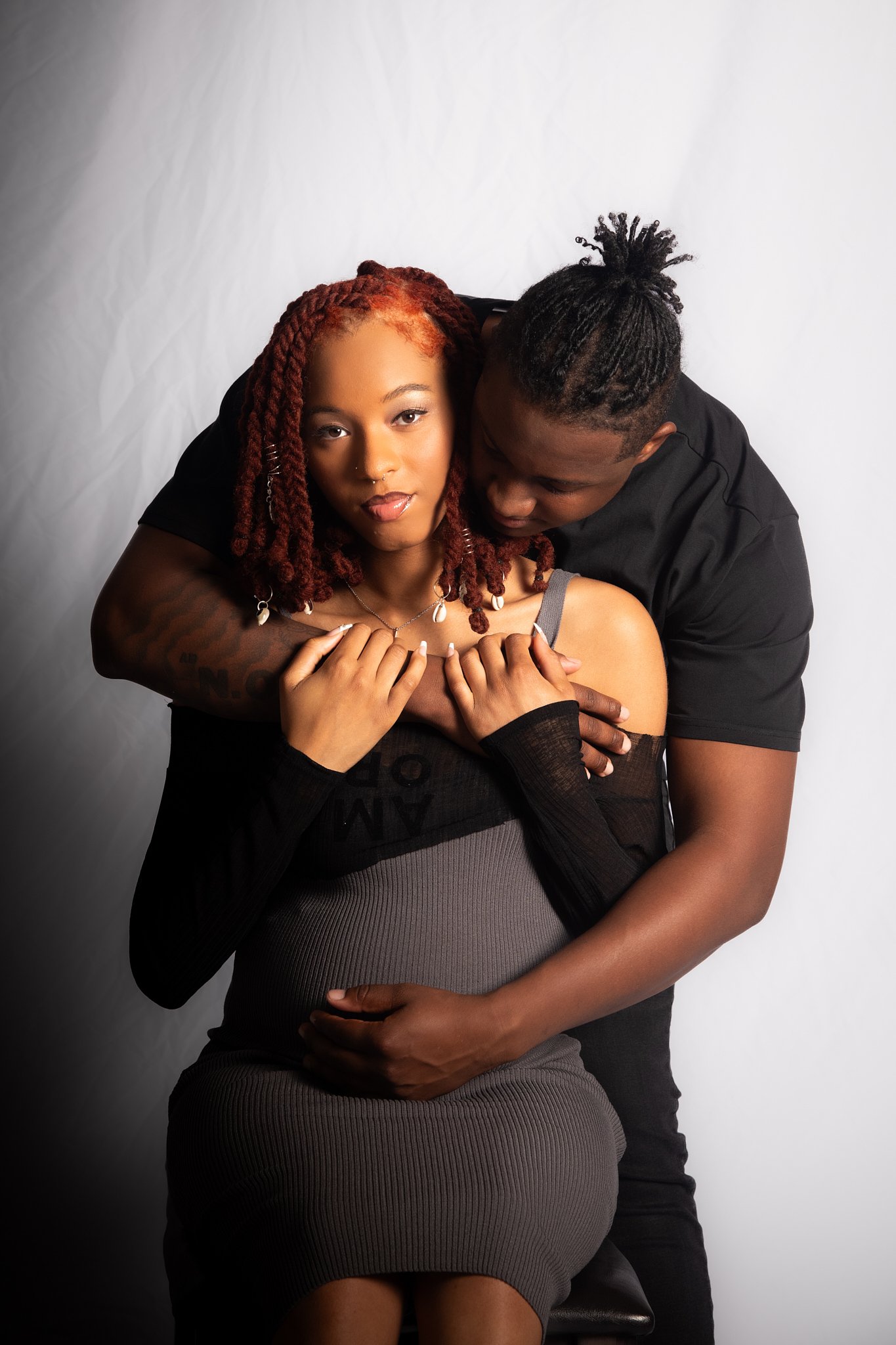 Pregnant woman sitting on a chair, embraced from behind by a man, against a neutral background.