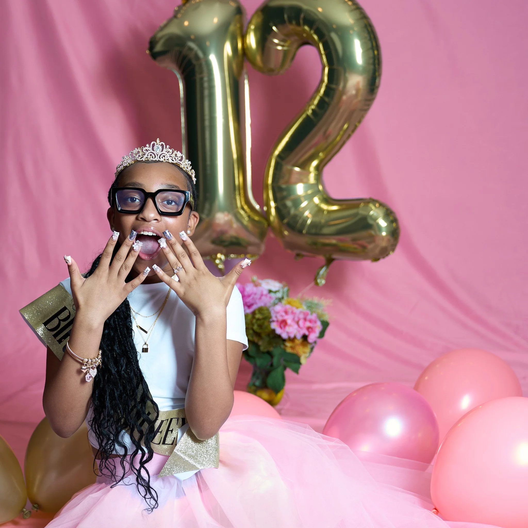 Girl celebrating 12th birthday with tiara, sash, balloons, and flowers, wearing a pink skirt and displaying her nails.