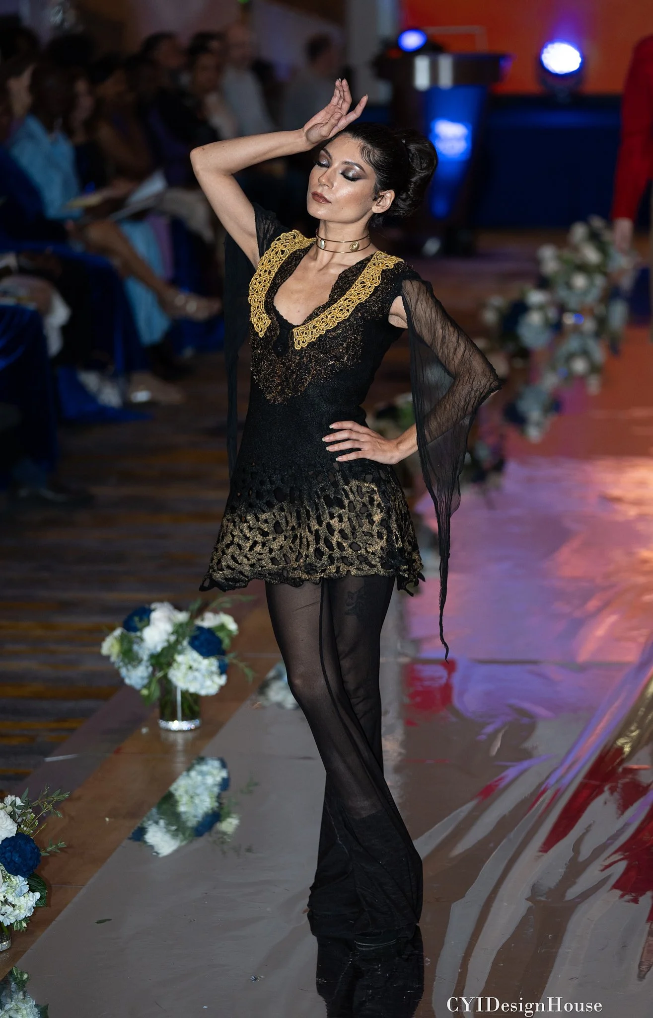 Model wearing a black and gold outfit on a runway, with hand raised to head, surrounded by an audience, floral arrangements, and blue lighting.