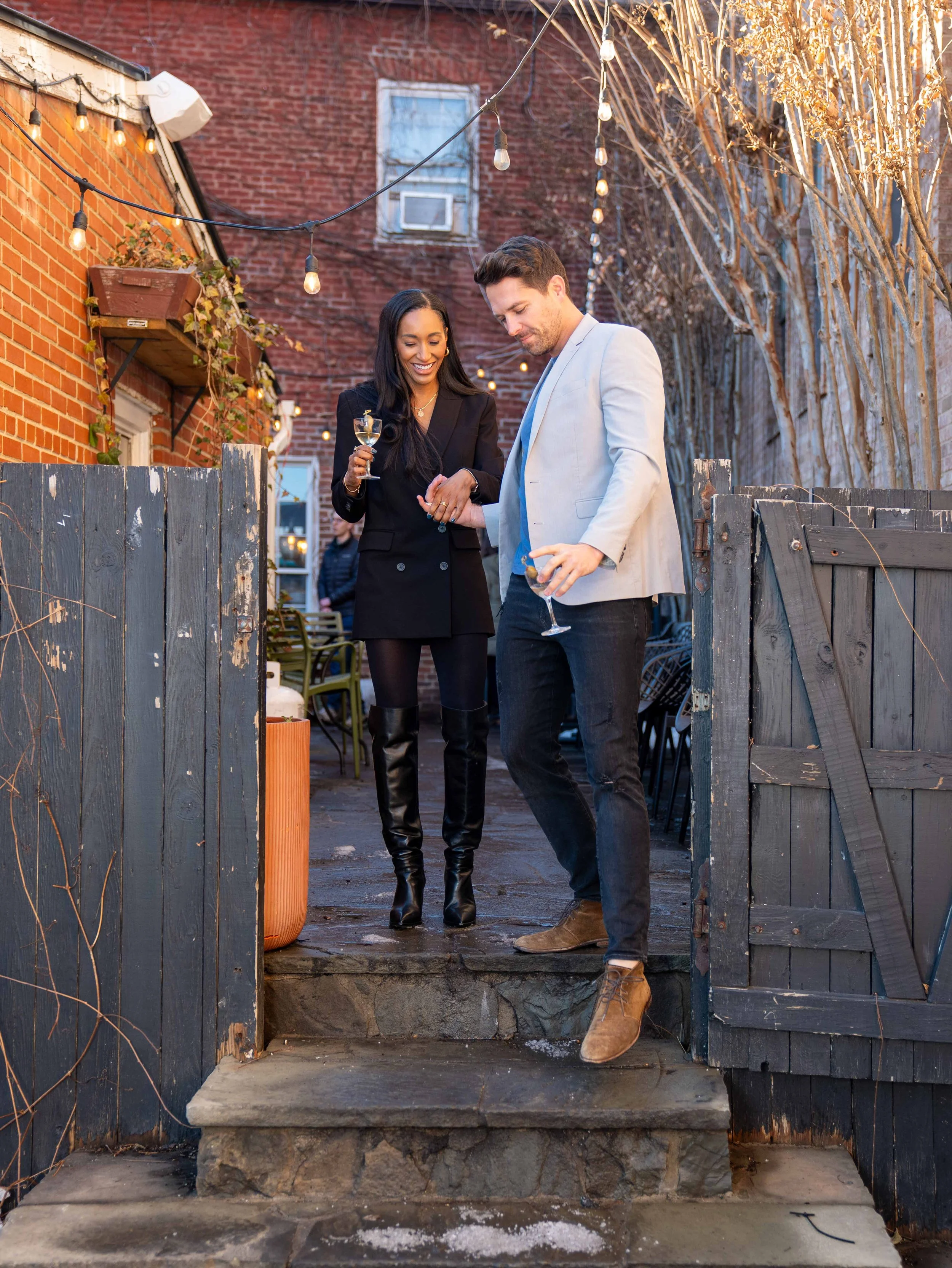 A smiling couple walking down outdoor steps, holding drinks, with a brick building and string lights in the background.