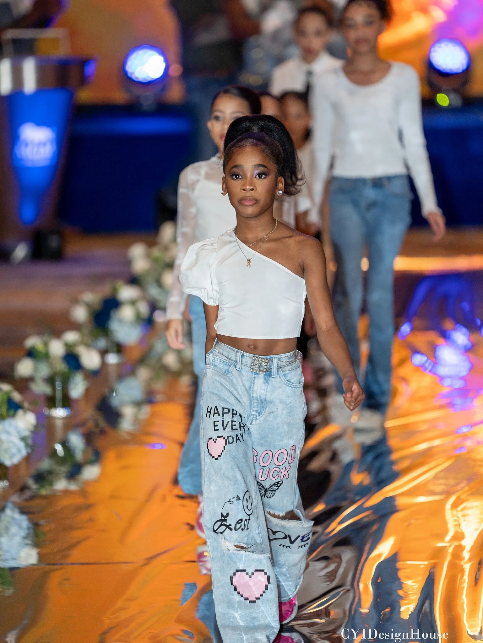 A young girl walks down a fashion runway wearing an asymmetric white top and light blue jeans with decorative patches and phrases. Other children in similar outfits follow behind her. The runway is reflective, and there are flowers on the sides. Blue