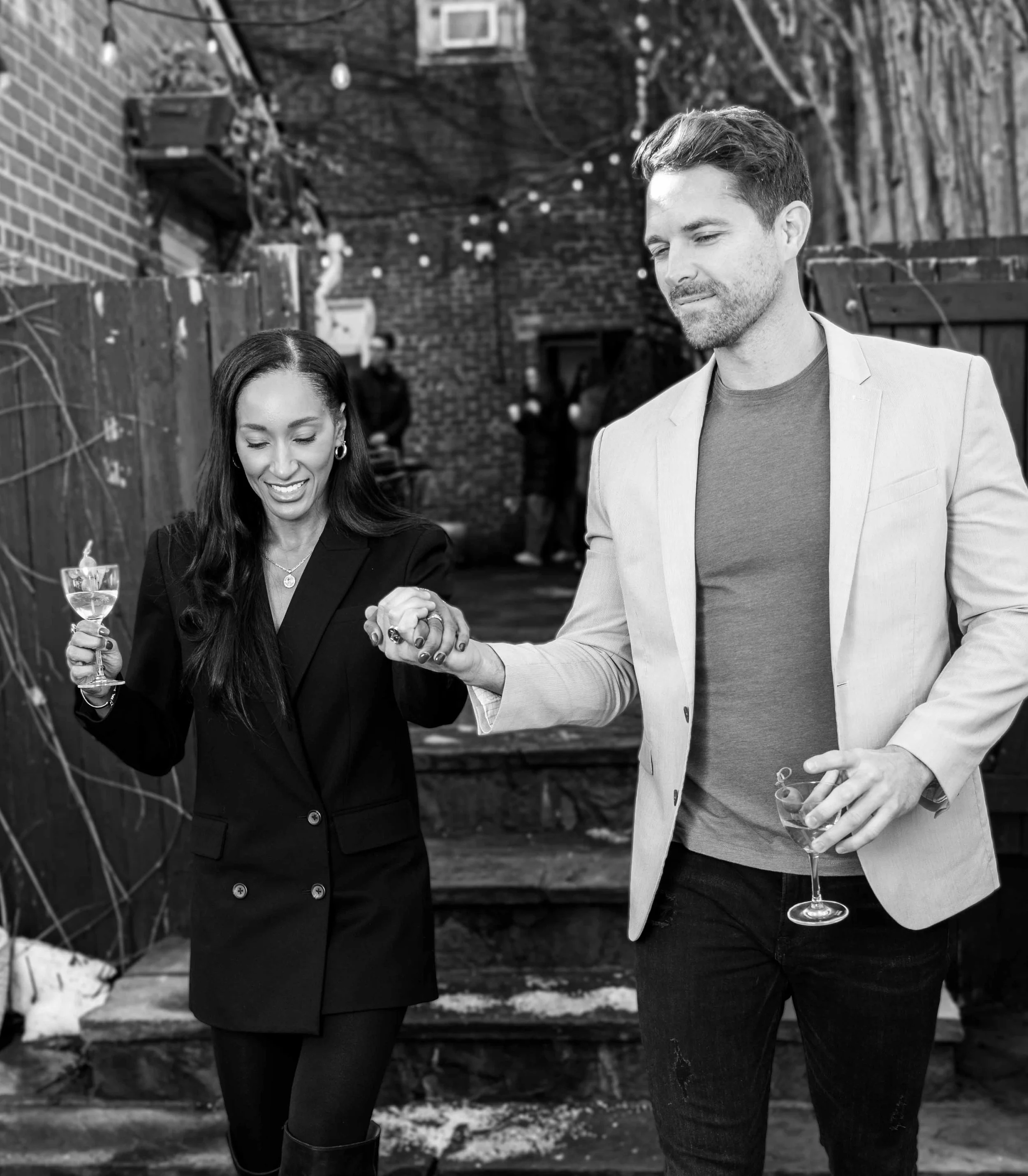 A man and woman holding hands while walking down steps and holding wine glasses, in a courtyard with string lights.