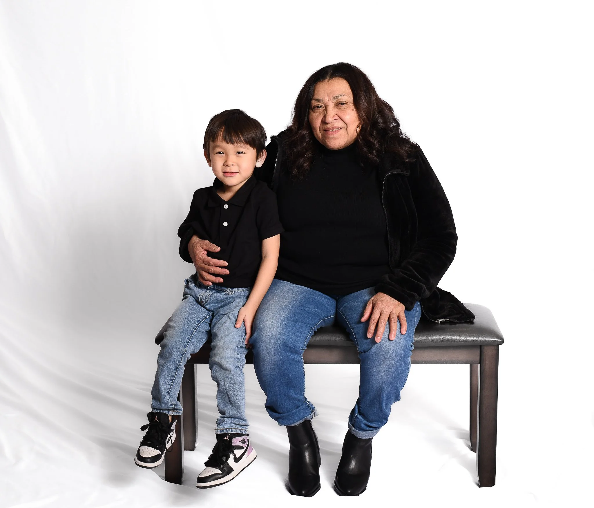 A young boy and an older woman sitting together on a bench against a white background. They are both wearing black tops and jeans.