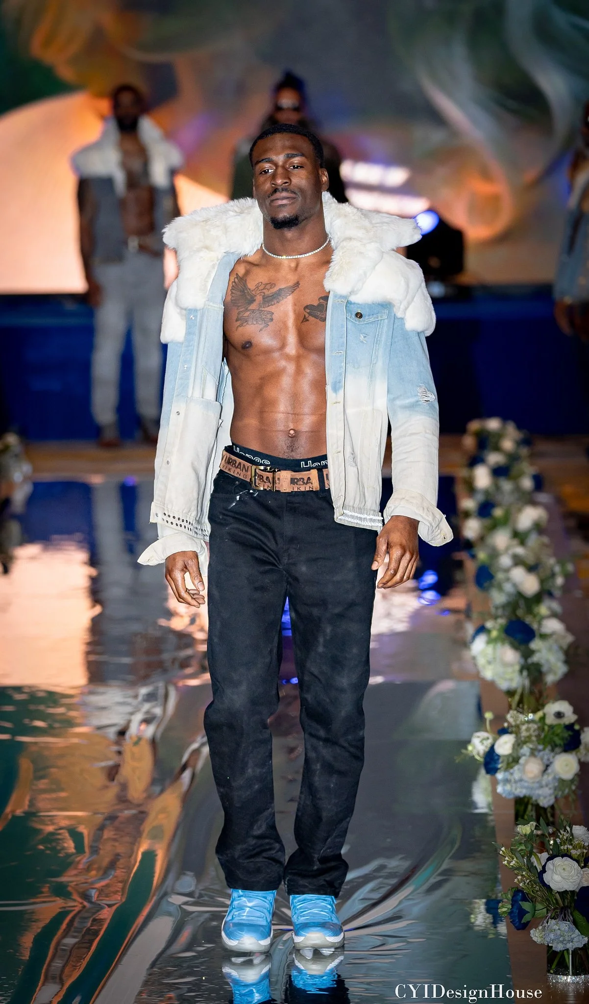 Male model walking on a fashion runway wearing a denim jacket with fur trim and black jeans, surrounded by floral decorations.