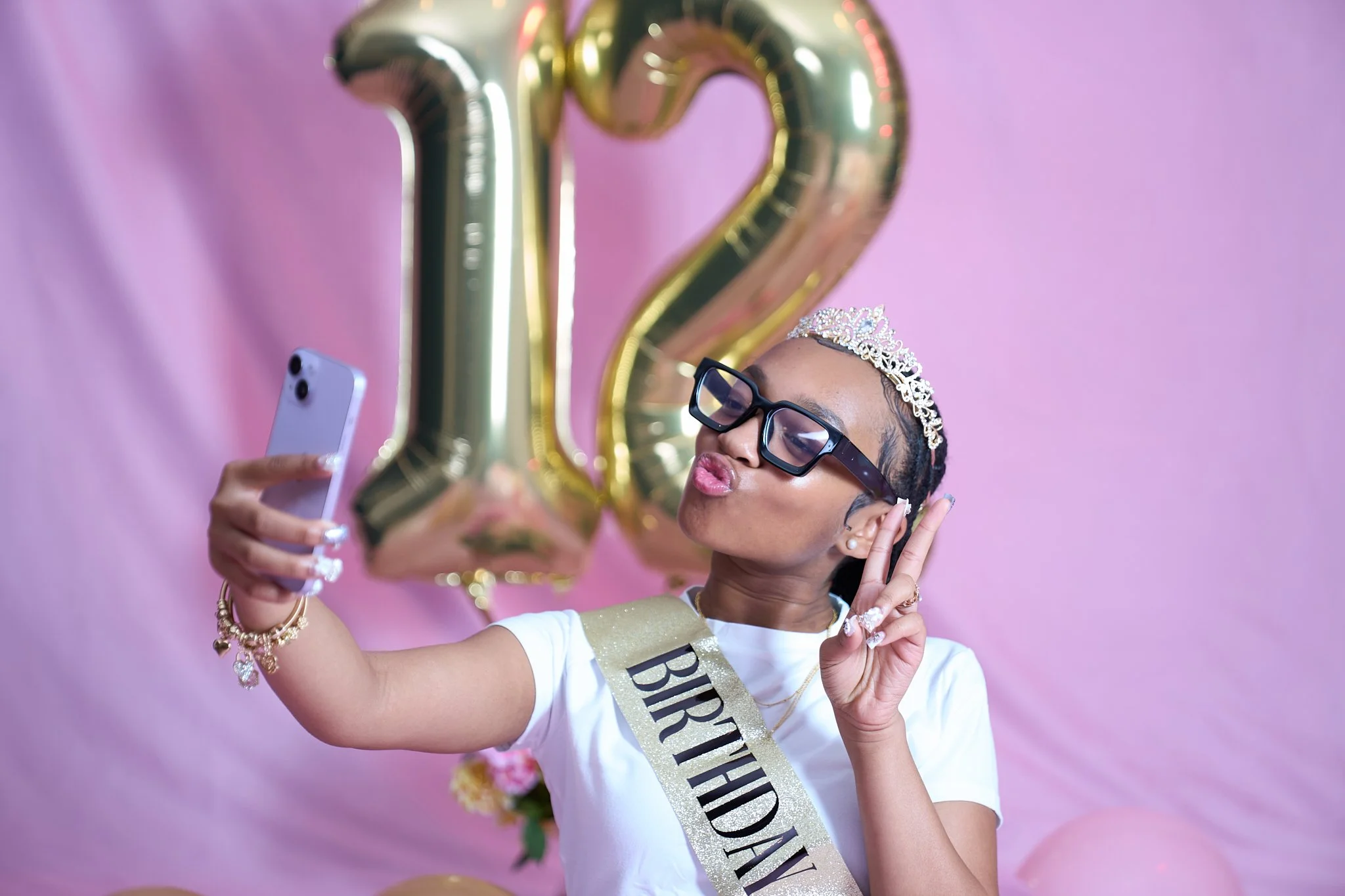Girl taking a selfie with a phone, wearing a tiara and glasses, next to golden "12" balloons and a "Birthday" sash, against a pink background.