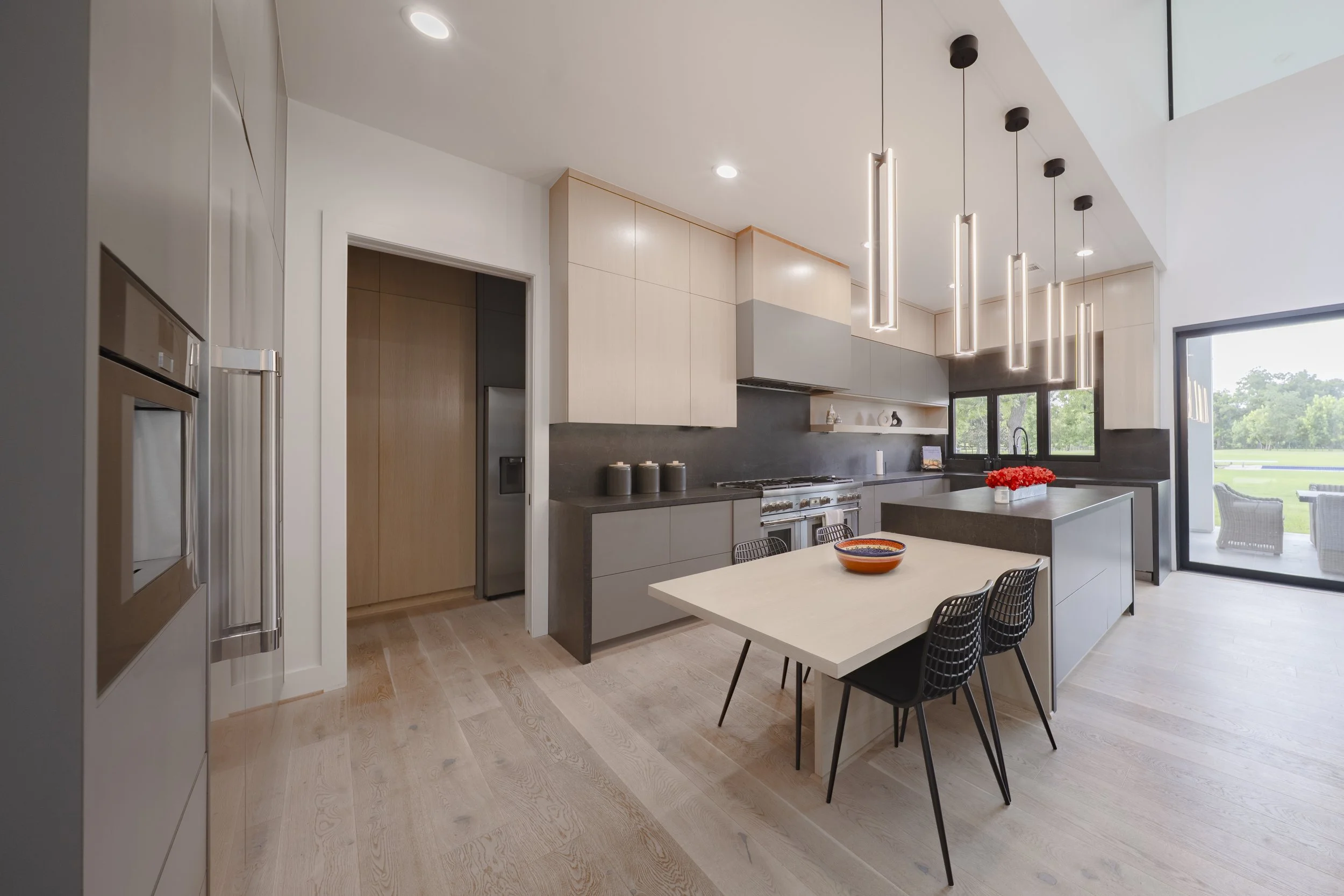 Modern kitchen with light wood cabinets, stainless steel appliances, black backsplash, and a large window overlooking a green outdoor area. Features a white island table with black chairs and pendant lighting.