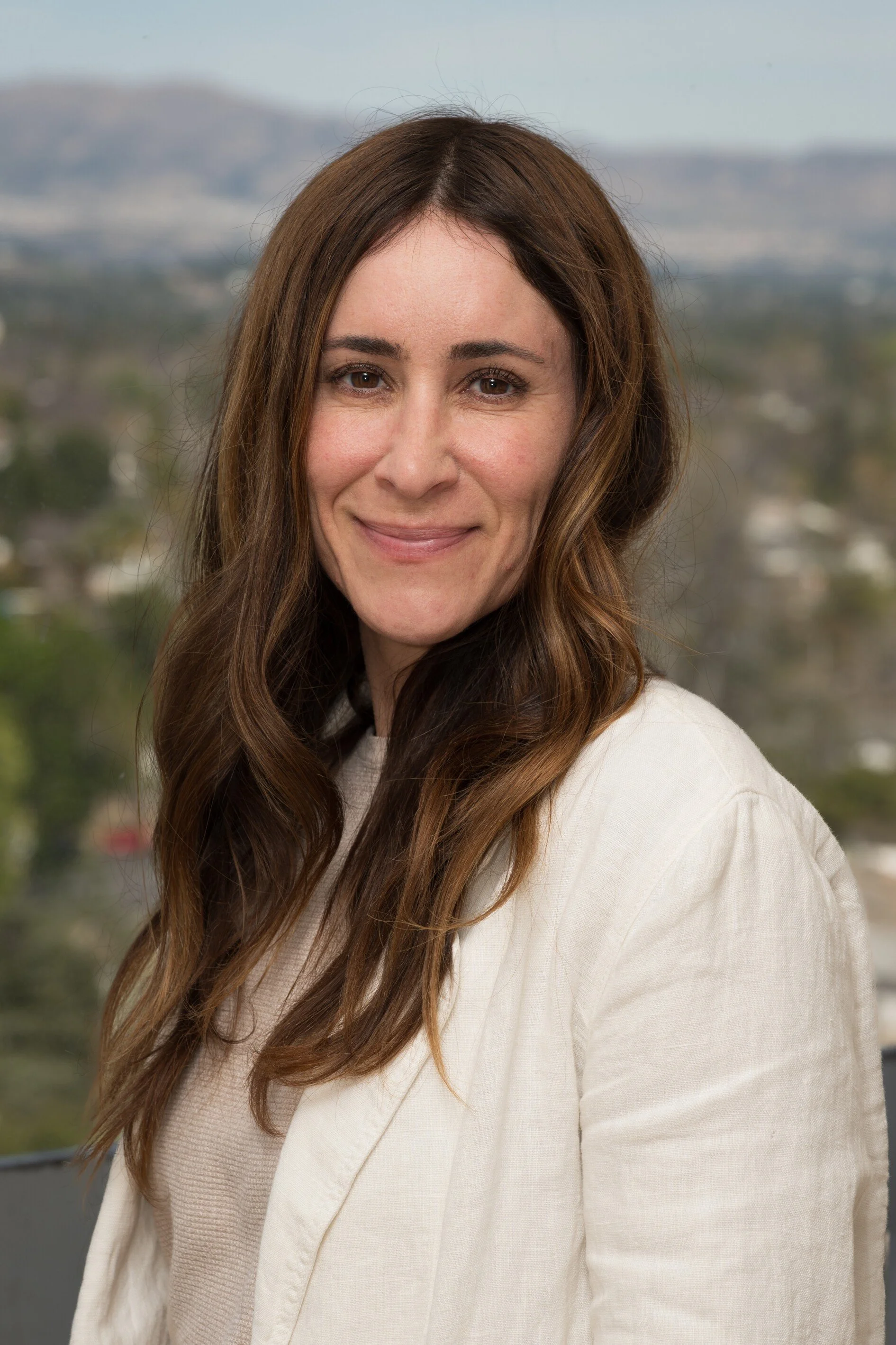 Portrait of a beautiful woman with long brown hair, wearing a light-colored jacket, smiling outdoors with a blurred natural landscape in the background.