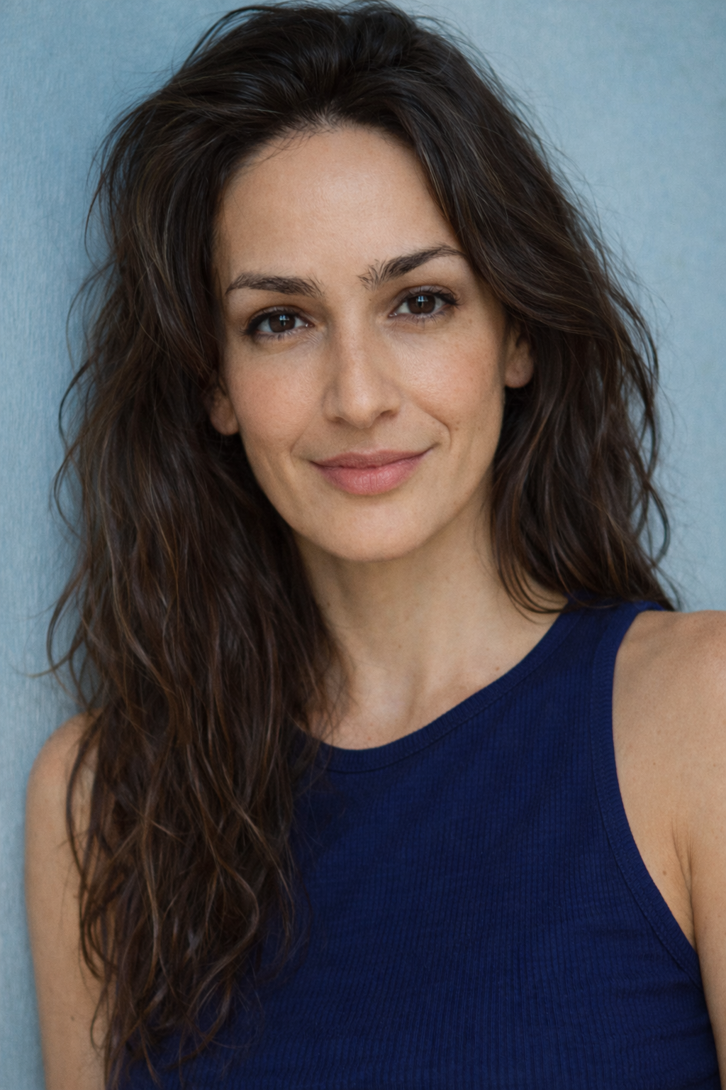 A woman with long, wavy brown hair and light skin, smiling at the camera, wearing a navy blue sleeveless top, against a light blue background.