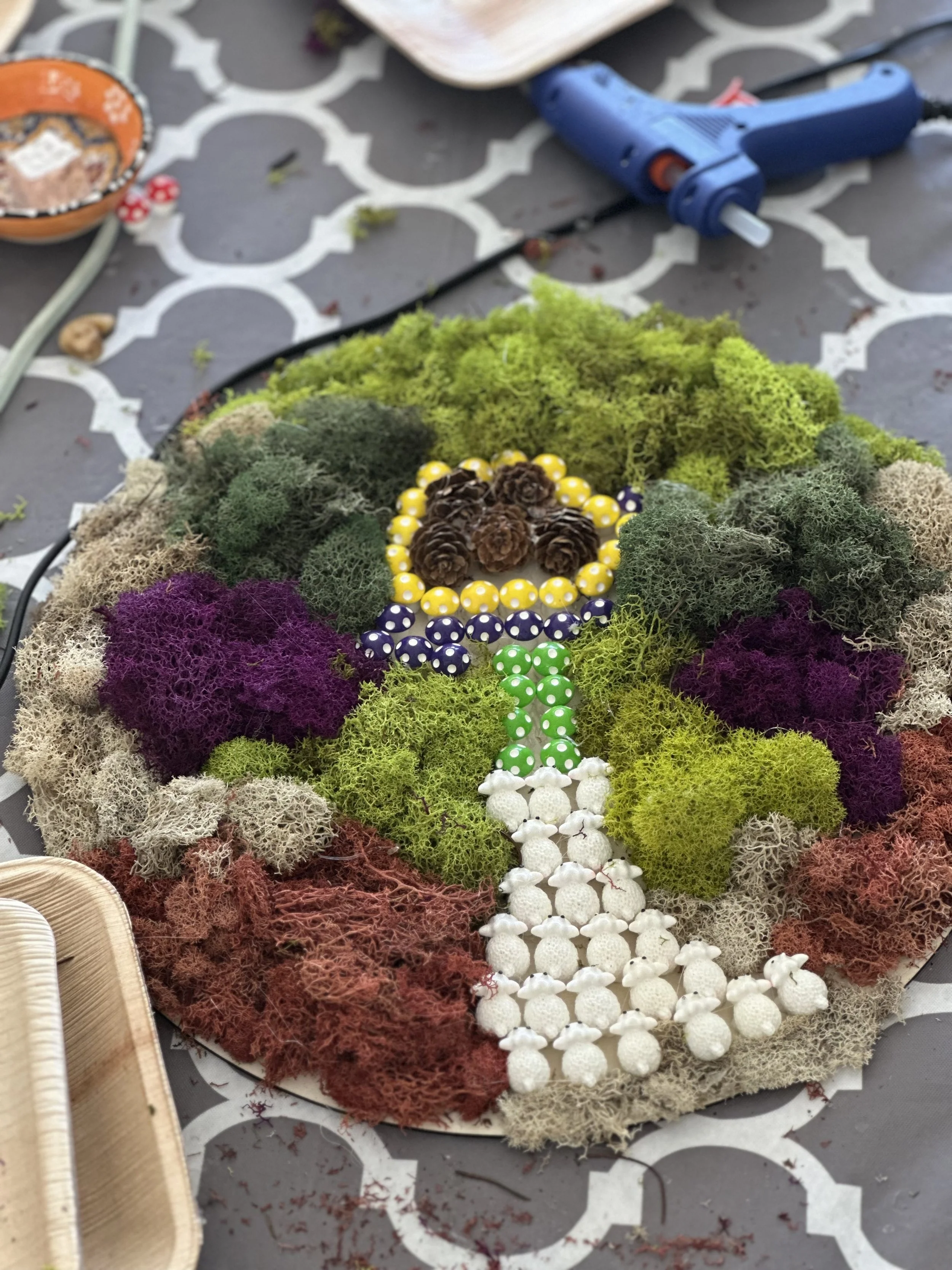Colorful moss arrangement with pinecones in the center, decorated with yellow, blue, and green polka-dot beads, on a patterned table.