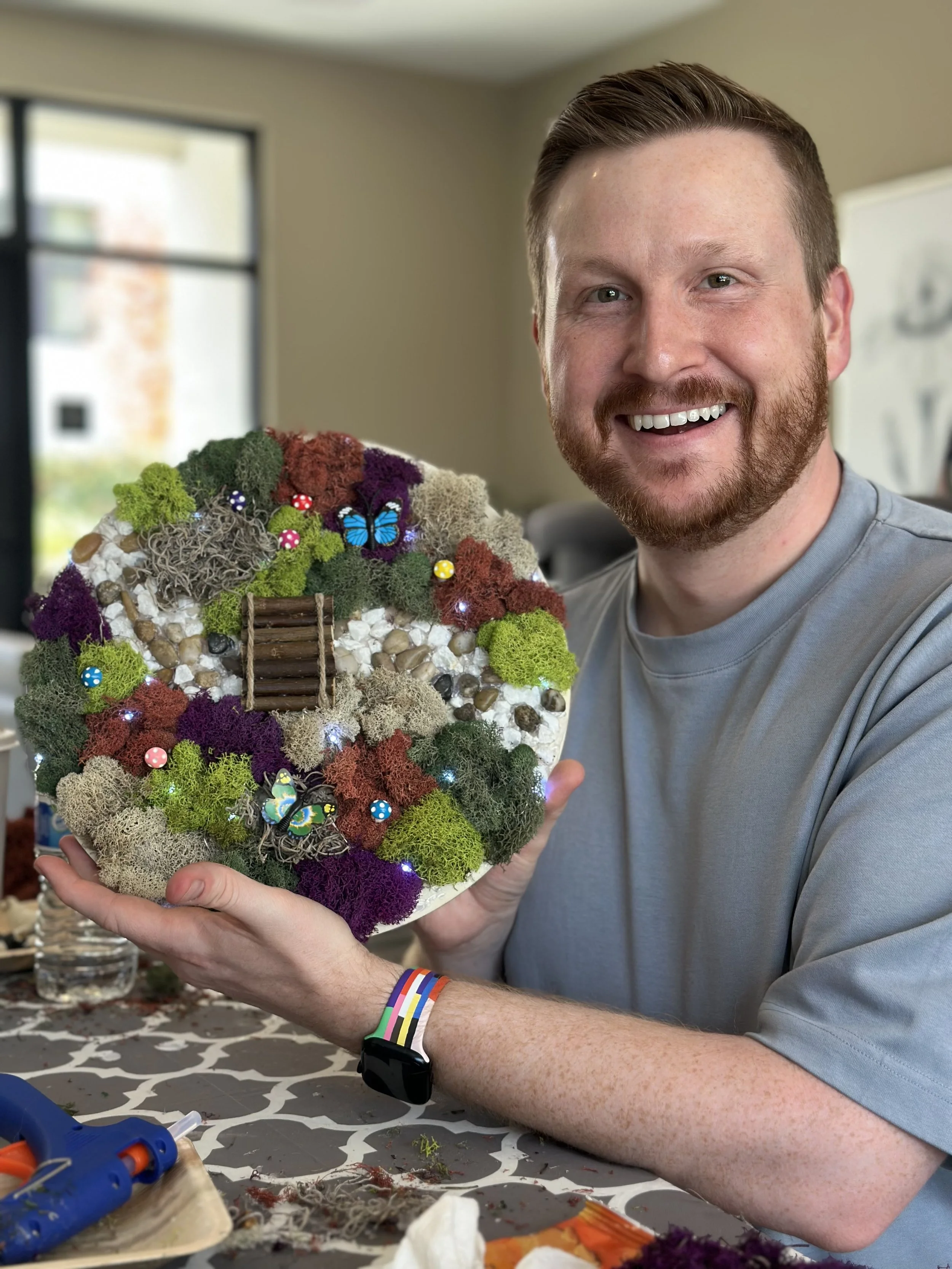 A man with a beard and short hair smiling and holding a decorative wreath made of moss, sticks, shells, and small ornaments inside a room with a window in the background.